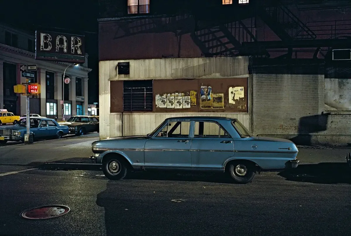 A vintage blue car parked on a city street at night, with a bar and additional vehicles visible in the background.