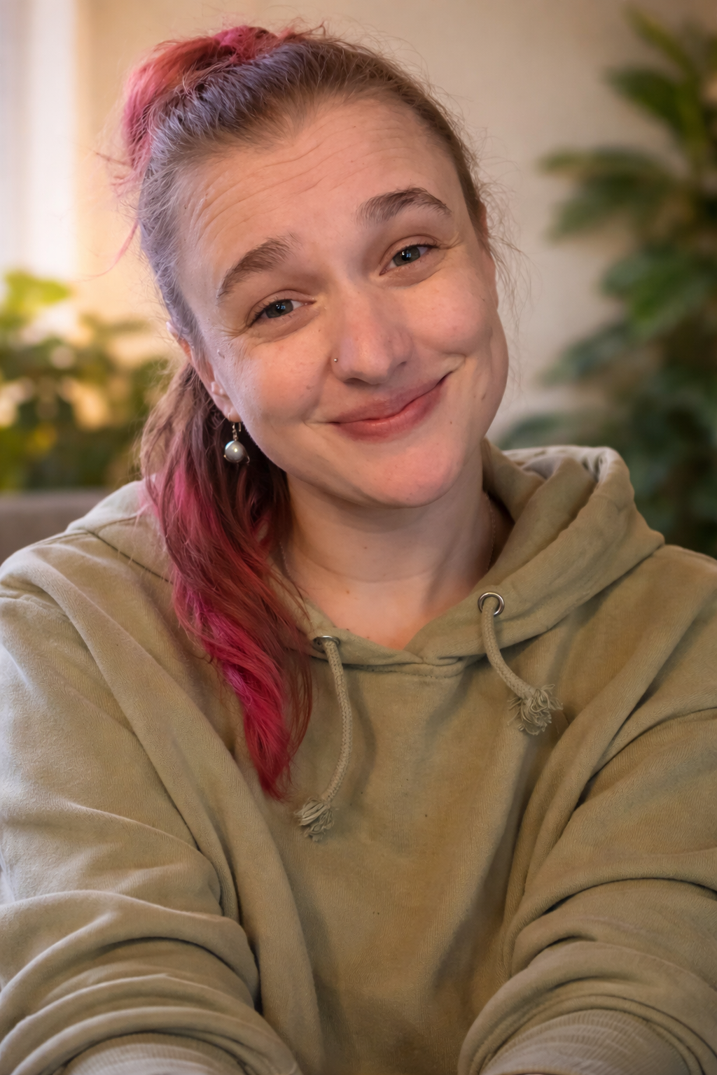 A smiling woman with pink hair, wearing a beige hoodie and pearl earrings, sitting indoors with greenery in the background.