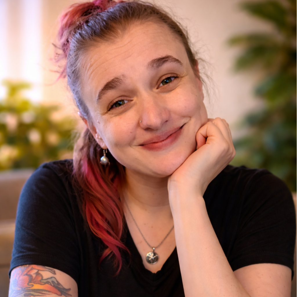 A woman with red hair and a small nose piercing, smiling and resting her chin on her hand, wearing a black shirt, jewelry, and earrings, with green plants in the background.
