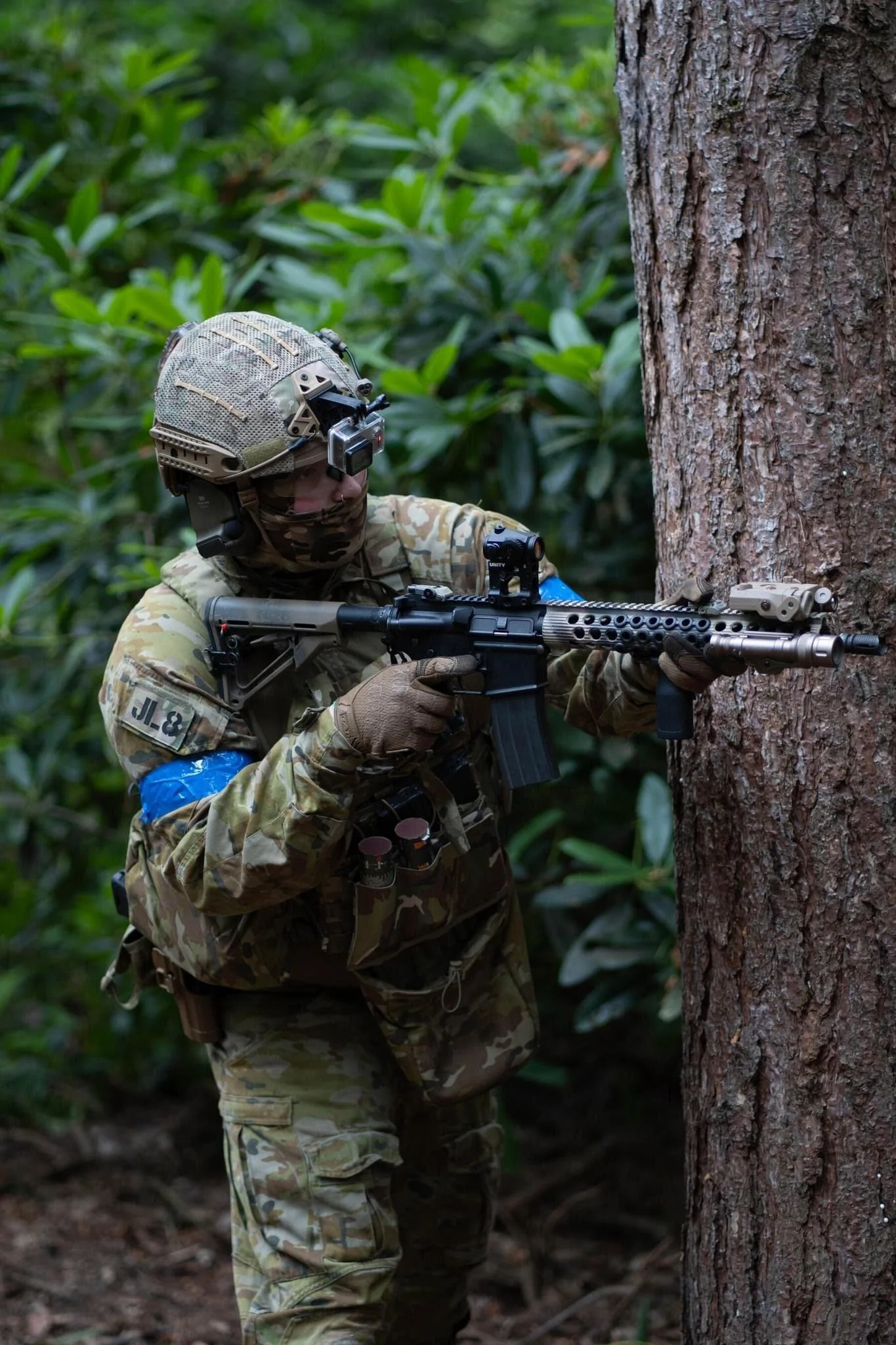 A soldier in camouflage gear aiming a rifle with a silencer in a forested area.