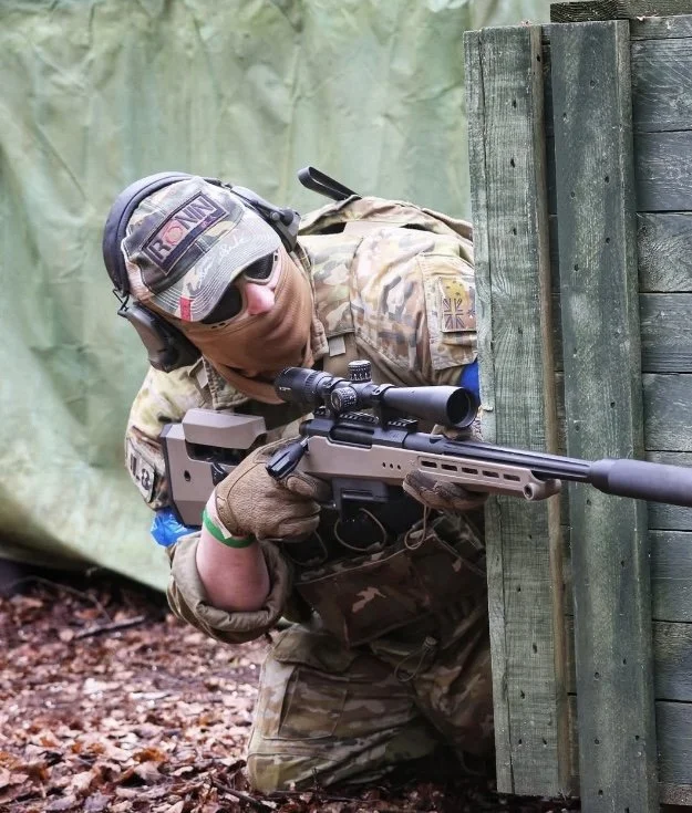 A soldier in camouflage uniform aiming a sniper rifle from behind a wooden barrier during a training exercise or military operation.
