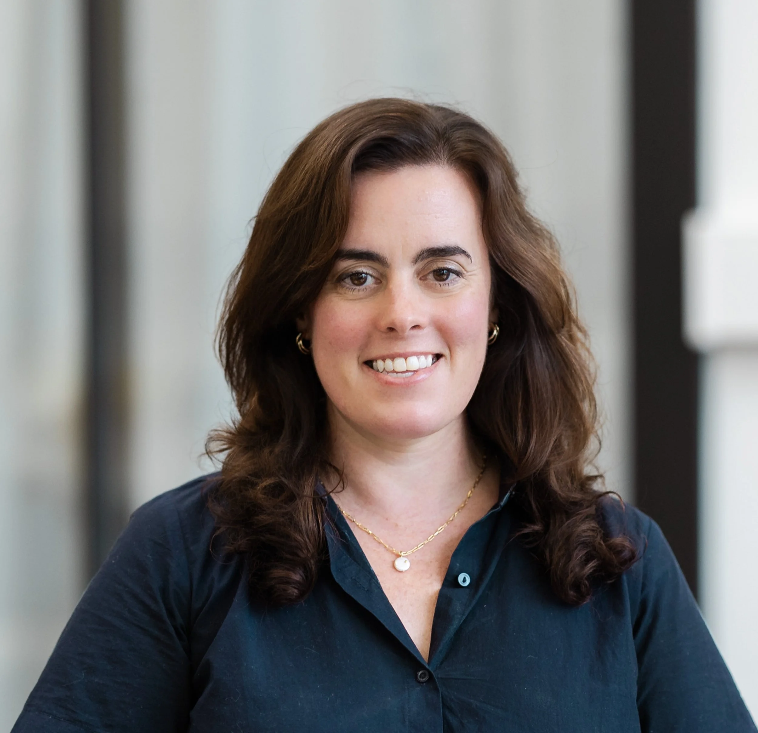 Portrait of a woman with shoulder-length brown hair, smiling, wearing a dark blue shirt, gold earrings, and a gold necklace with a pearl pendant.
