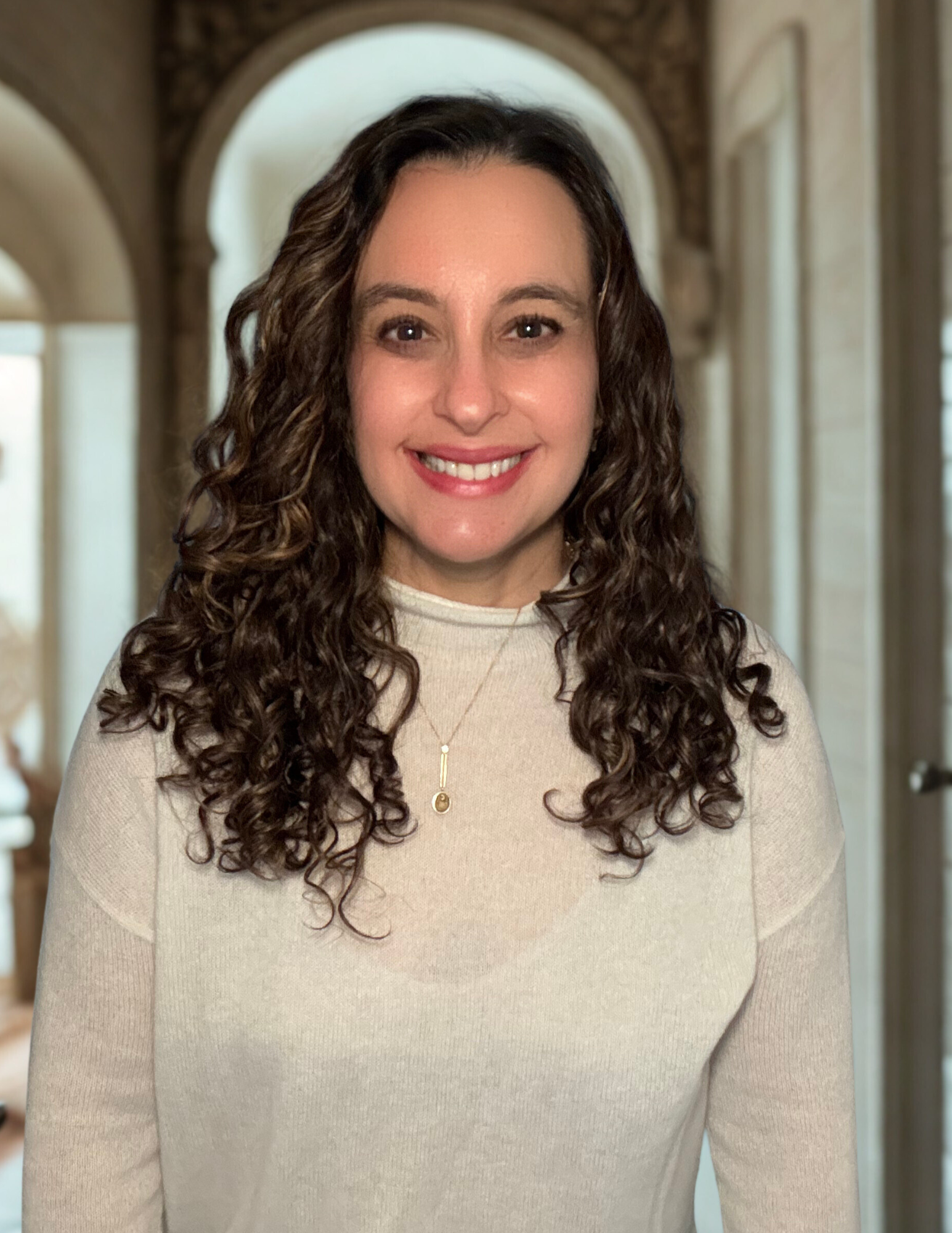 A woman with dark, curly hair smiling at the camera, wearing a light-colored top and a necklace, standing in a well-lit hallway with arched doorways.