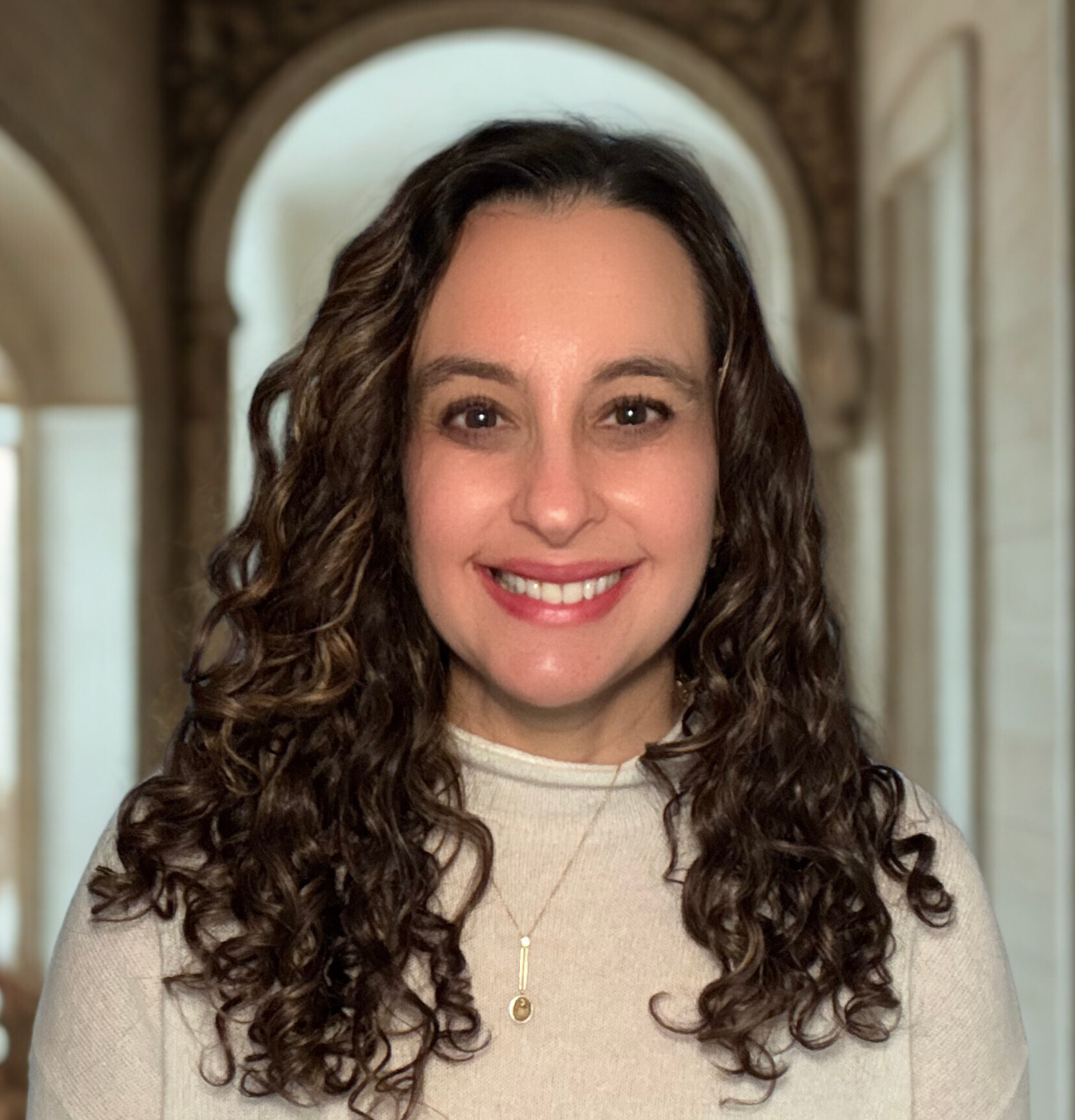 A woman with curly brown hair smiling, wearing a cream-colored top and a gold necklace, standing in a hallway with arched doorways.