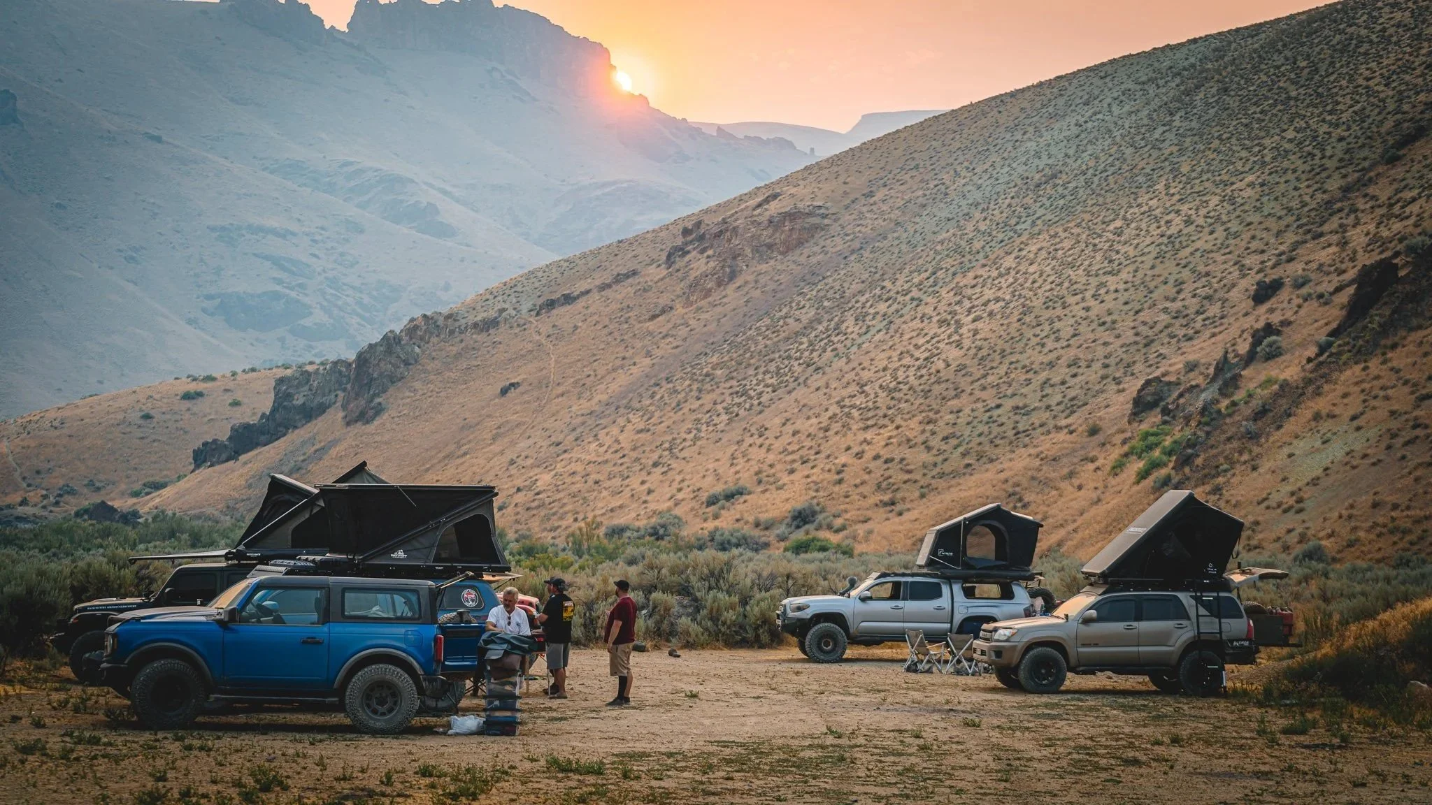 A group of people preparing for camping with three off-road vehicles equipped with rooftop tents in a desert landscape during sunset, with mountains in the background.