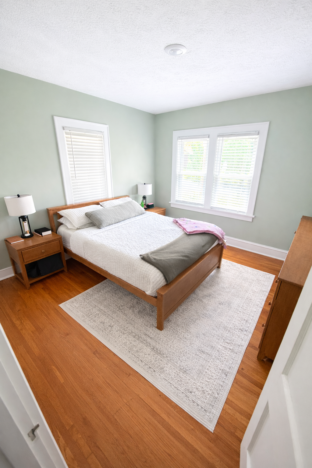 Room with a wooden bed frame, white bedding, two nightstands with lamps, a window with blinds, and a hardwood floor with an off-white area rug.