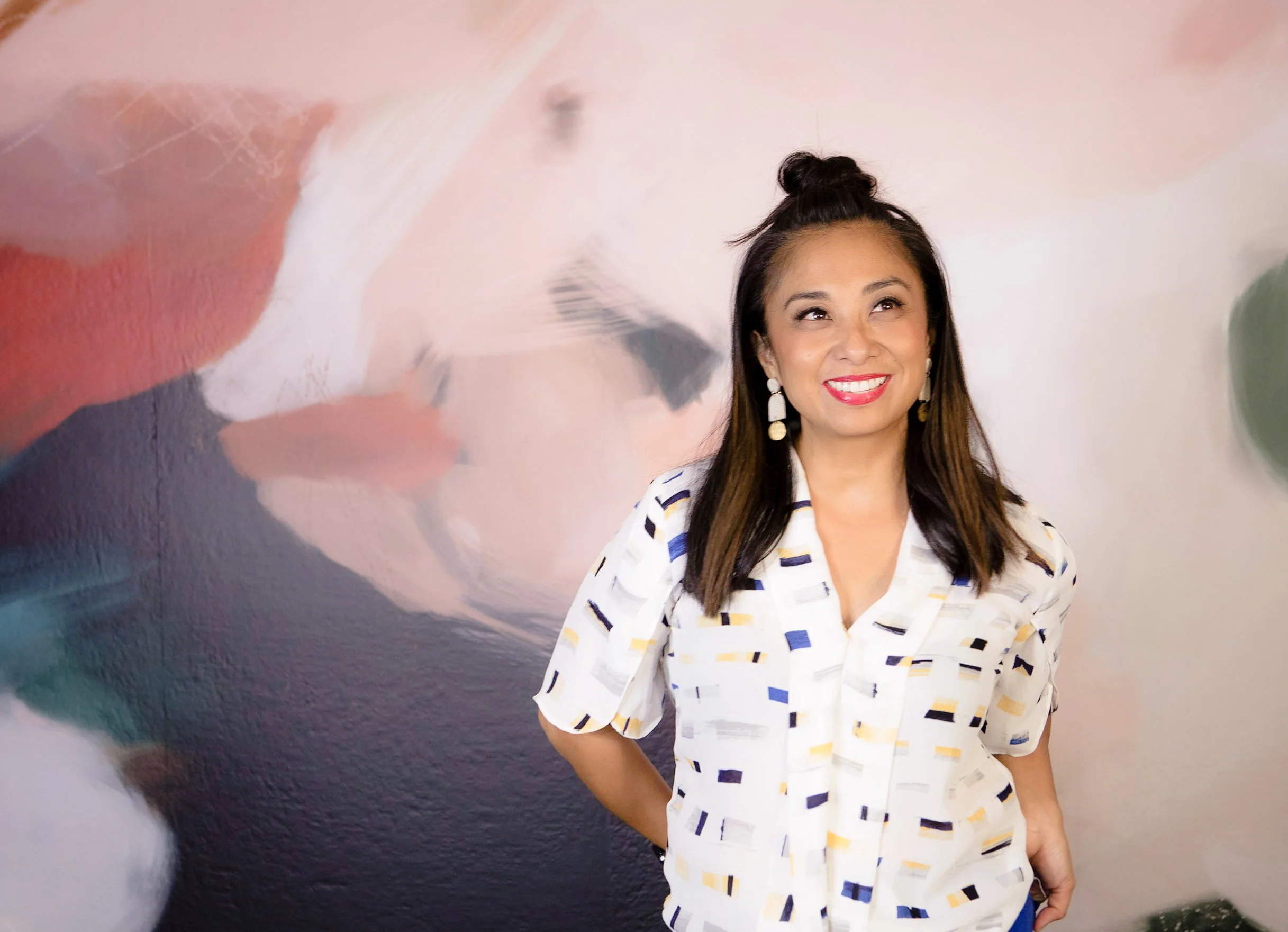 A woman with long brown hair wearing a white blouse with colorful block patterns, standing in front of an abstract pink, white, and dark mural background, smiling with bright red lipstick.