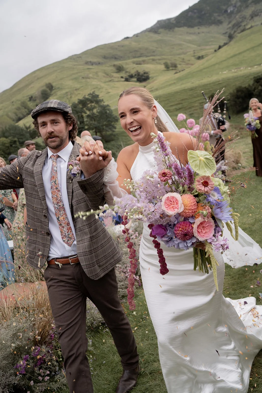 Newlyweds walking through a garden with a colourful bouquet and hills behind them.