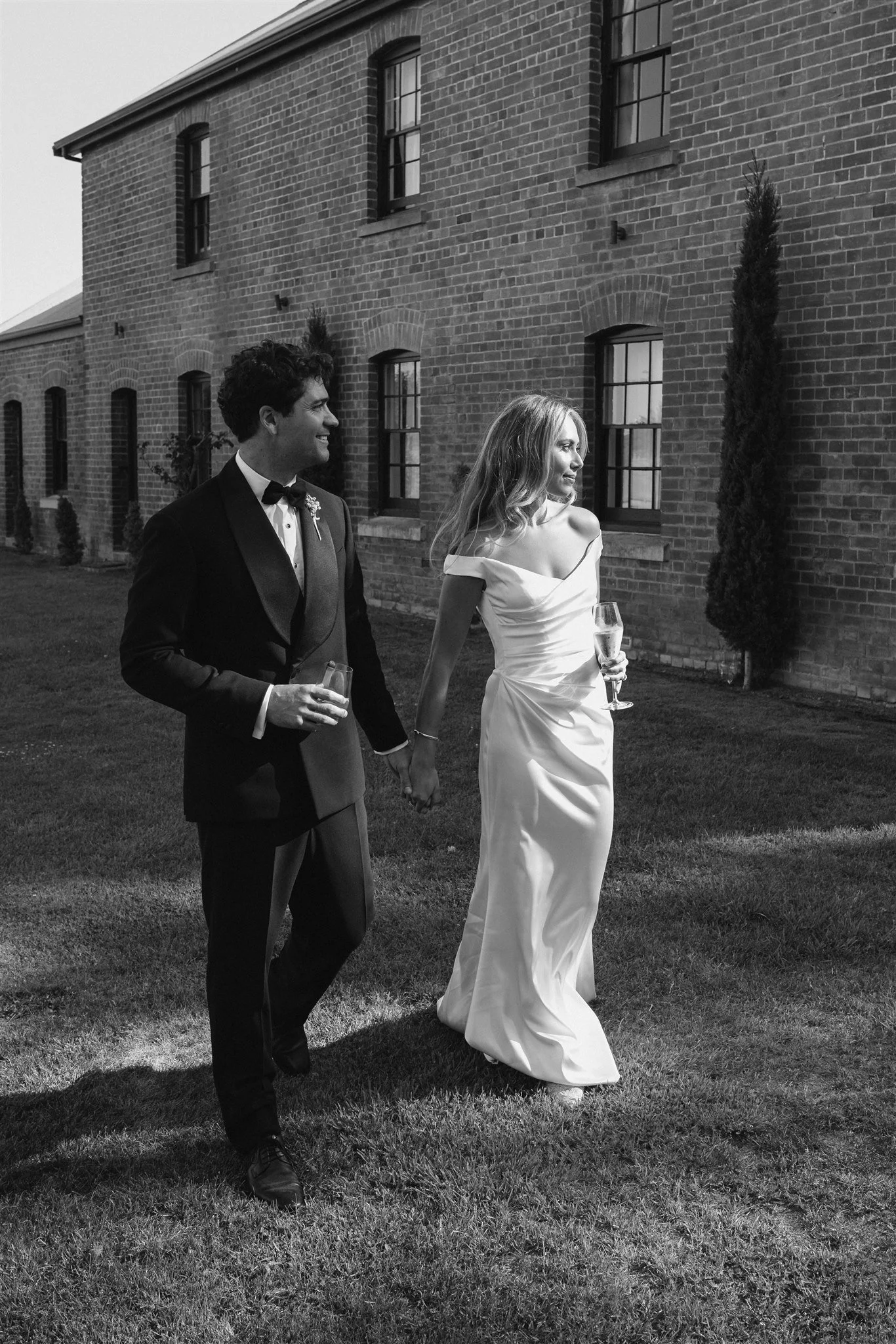 A black and white photograph of a couple dressed in wedding attire holding hands and walking outdoors on a grassy area, with a brick building and windows in the background. The man is wearing a tuxedo with a bow tie, and the woman is wearing a strapless wedding gown and holding a glass of champagne.