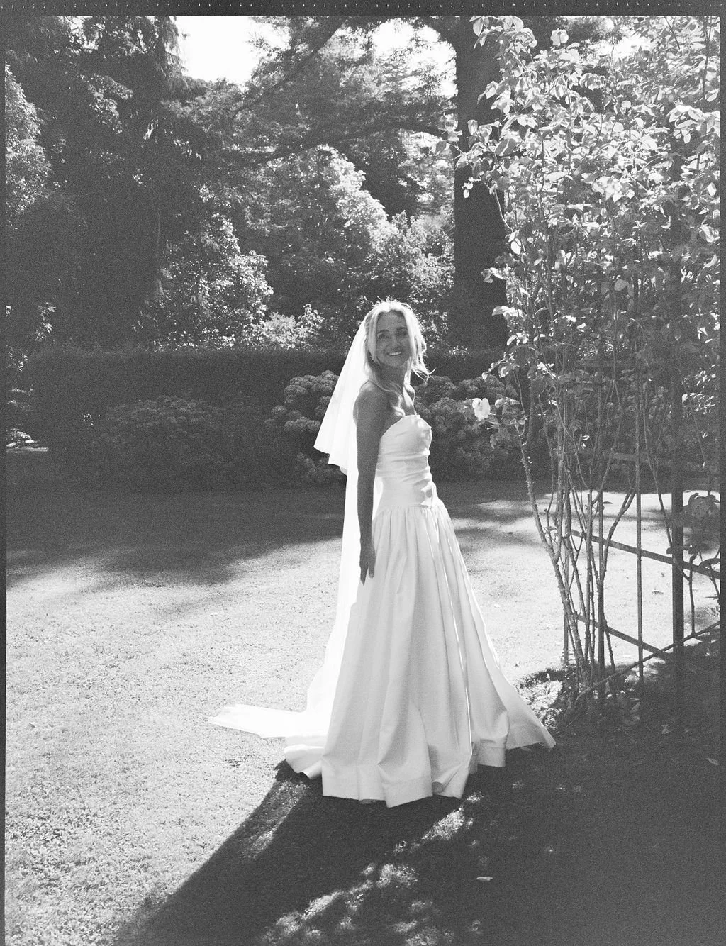 Black and white photo of a smiling bride standing outdoors near a vine-covered structure, in a lush garden with trees and shrubs in the background.