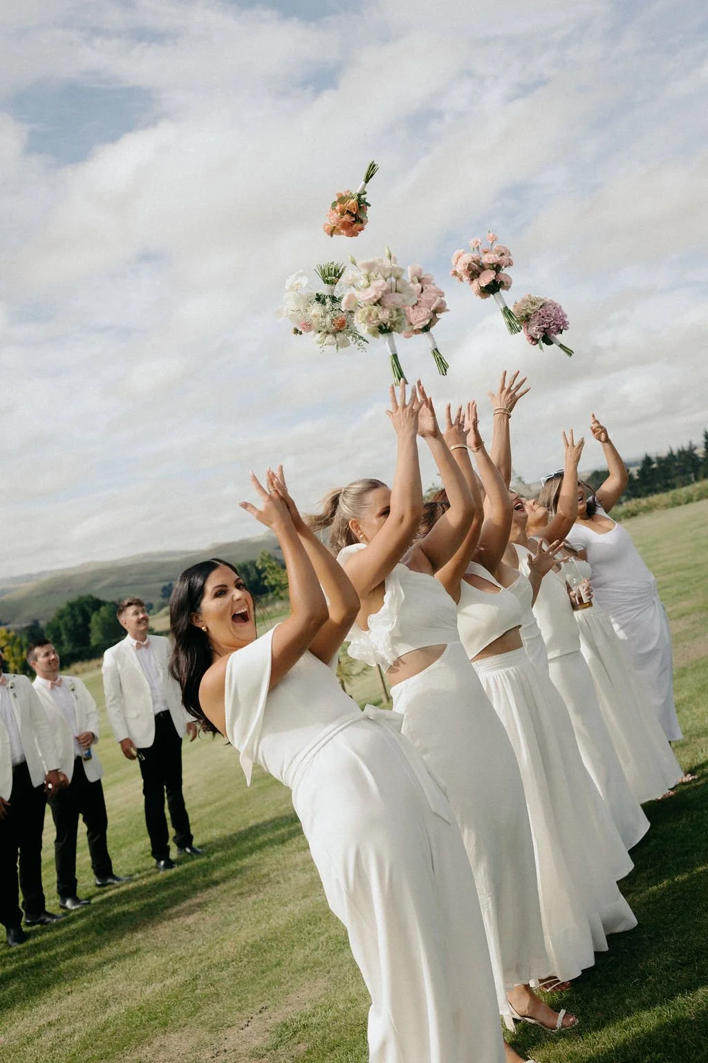 Bridesmaids tossing bouquets into the air at an outdoor wedding.