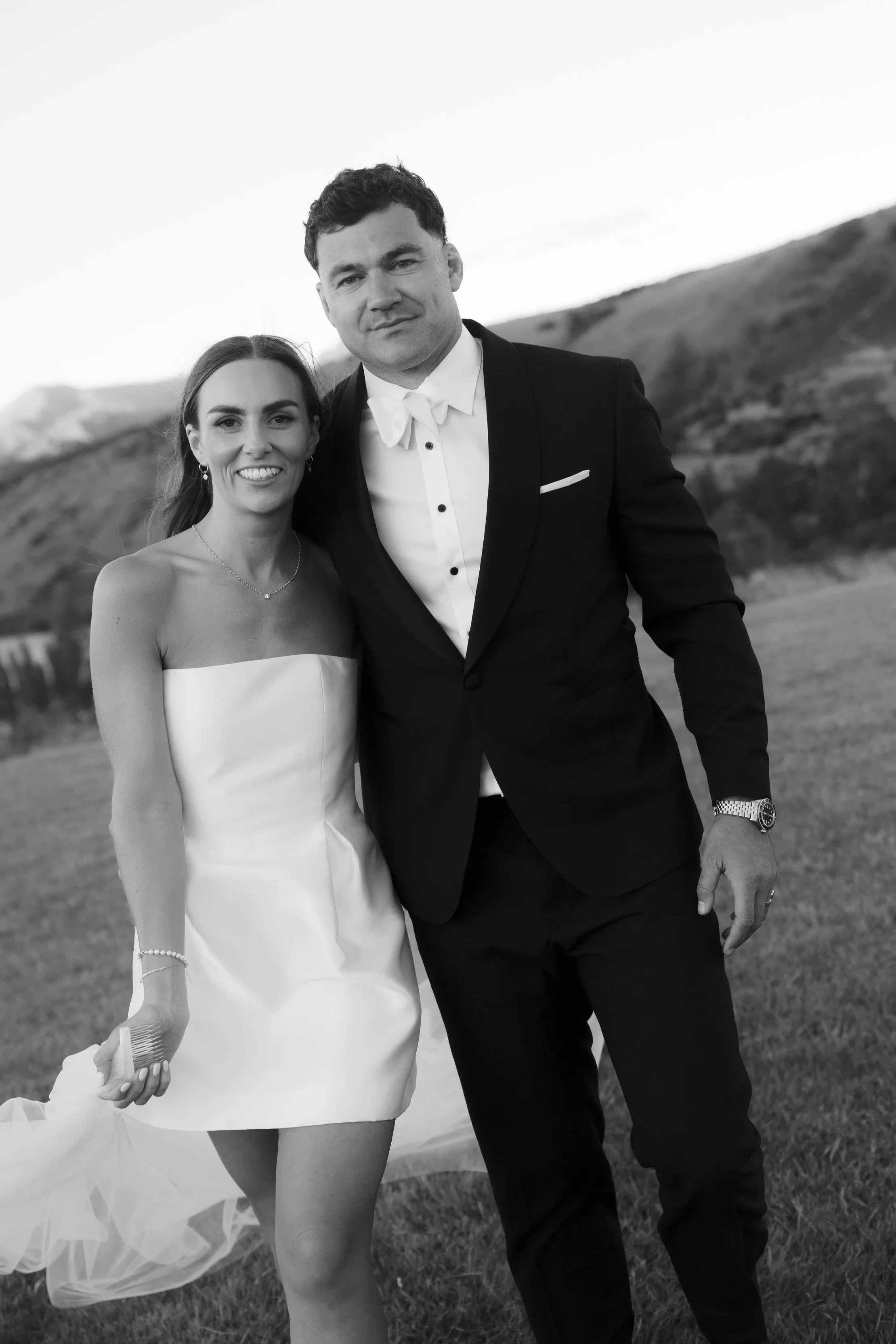 A black-and-white photo of a smiling woman in a strapless white dress and a man in a tuxedo, standing outdoors on a grassy field with hills in the background.