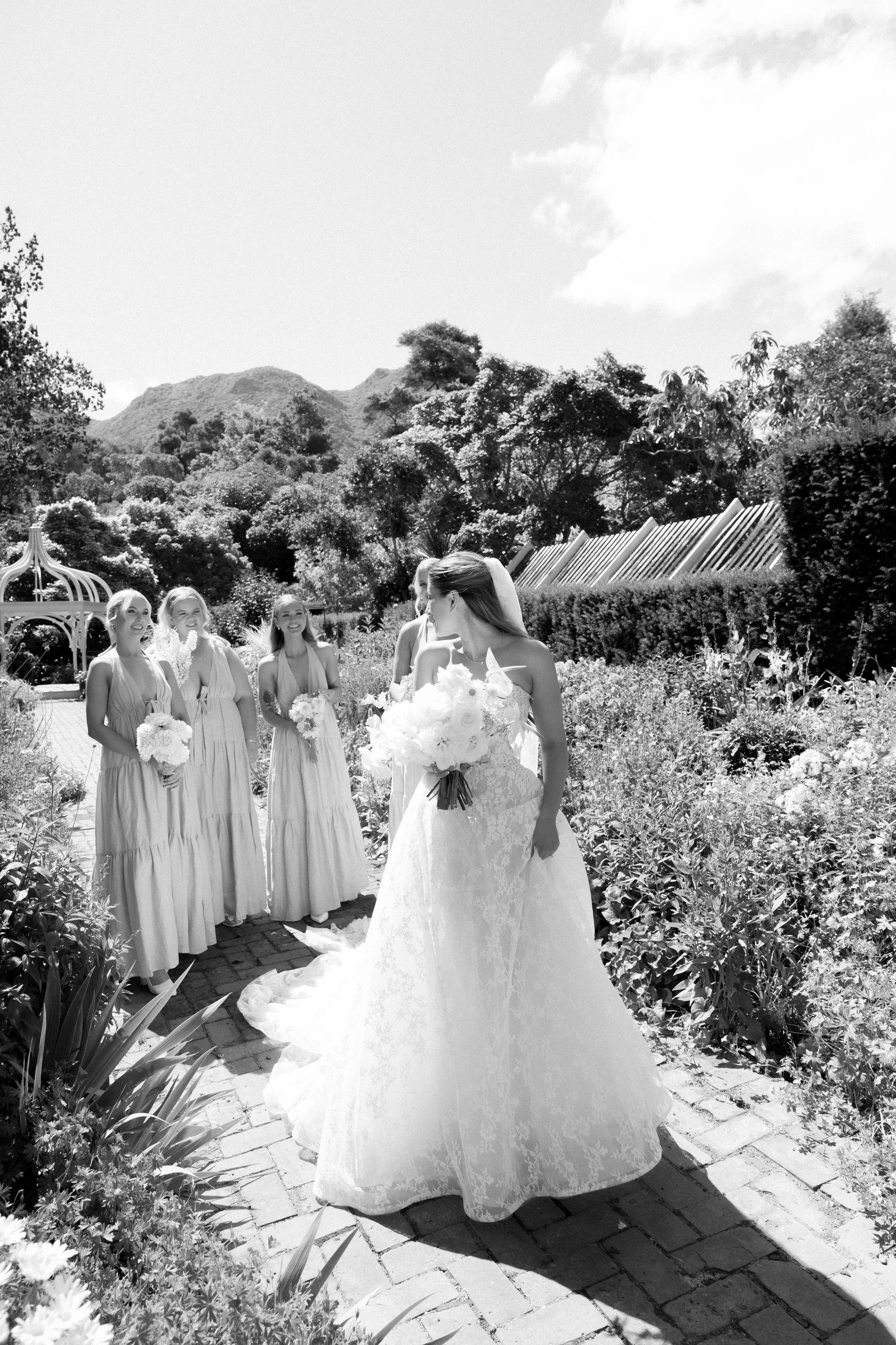 Bride and bridesmaids with bouquets walking through a garden, in black and white.
