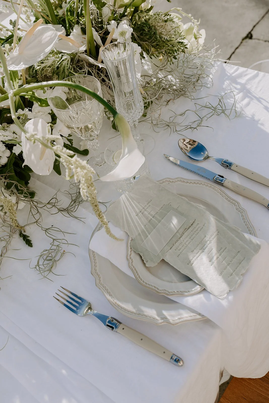 Elegant table setting with white plates, crystal glasses, silverware, and a floral centerpiece with white flowers and greenery on a white tablecloth.