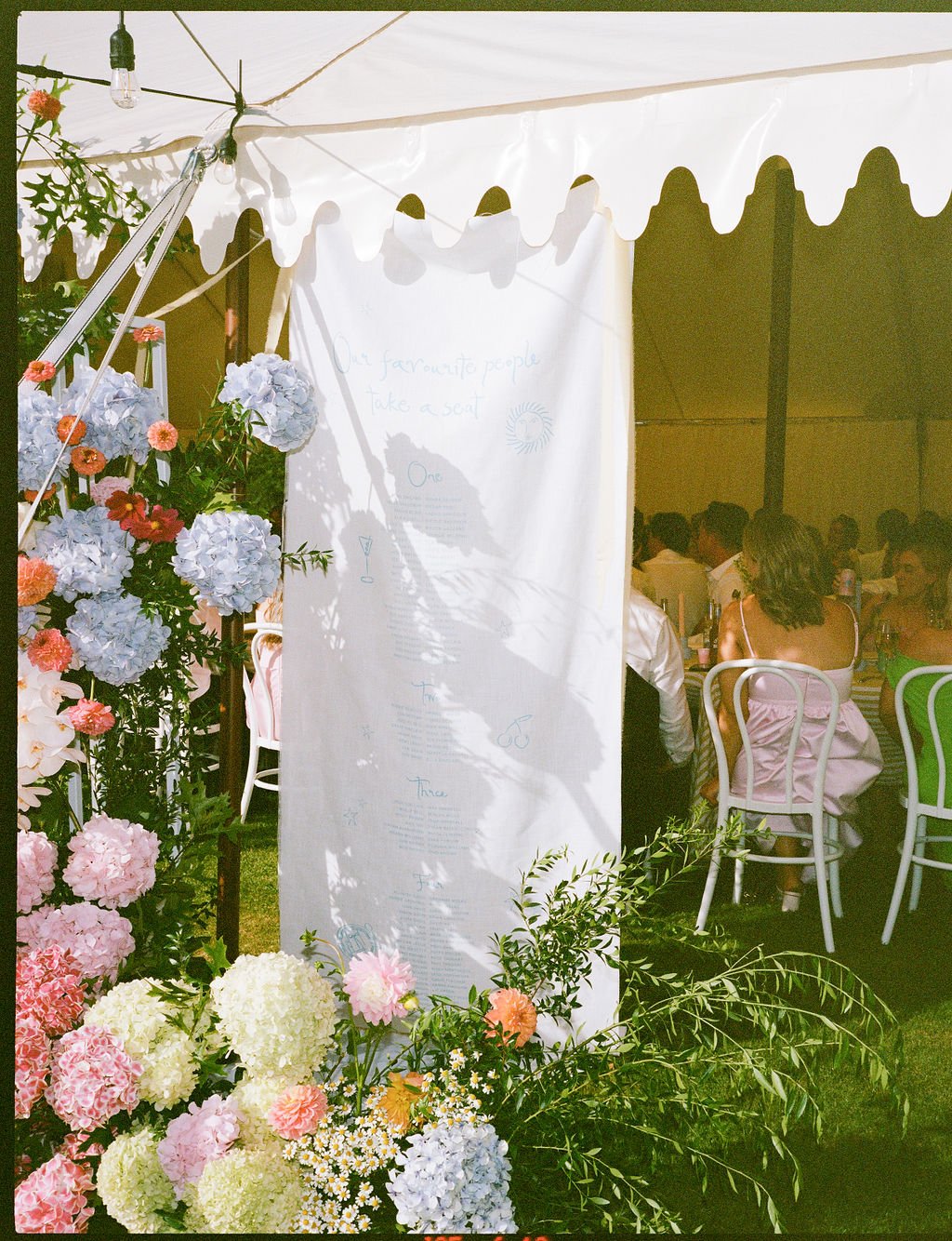 Outdoor event with a white tent, floral decorations, and guests seated at tables, with a whiteboard or sign hanging inside.