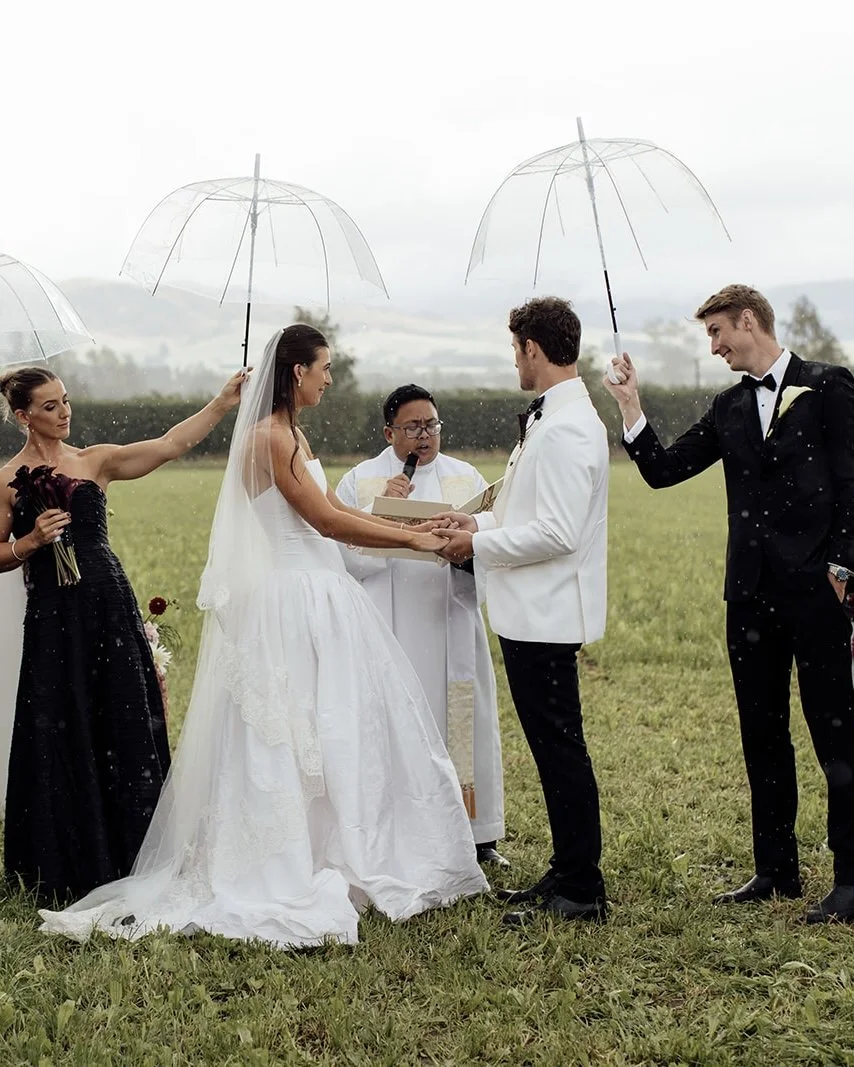 A wedding ceremony outdoors with a bride and groom holding hands, officiant, and bridesmaids and groomsmen holding umbrellas in light rain.