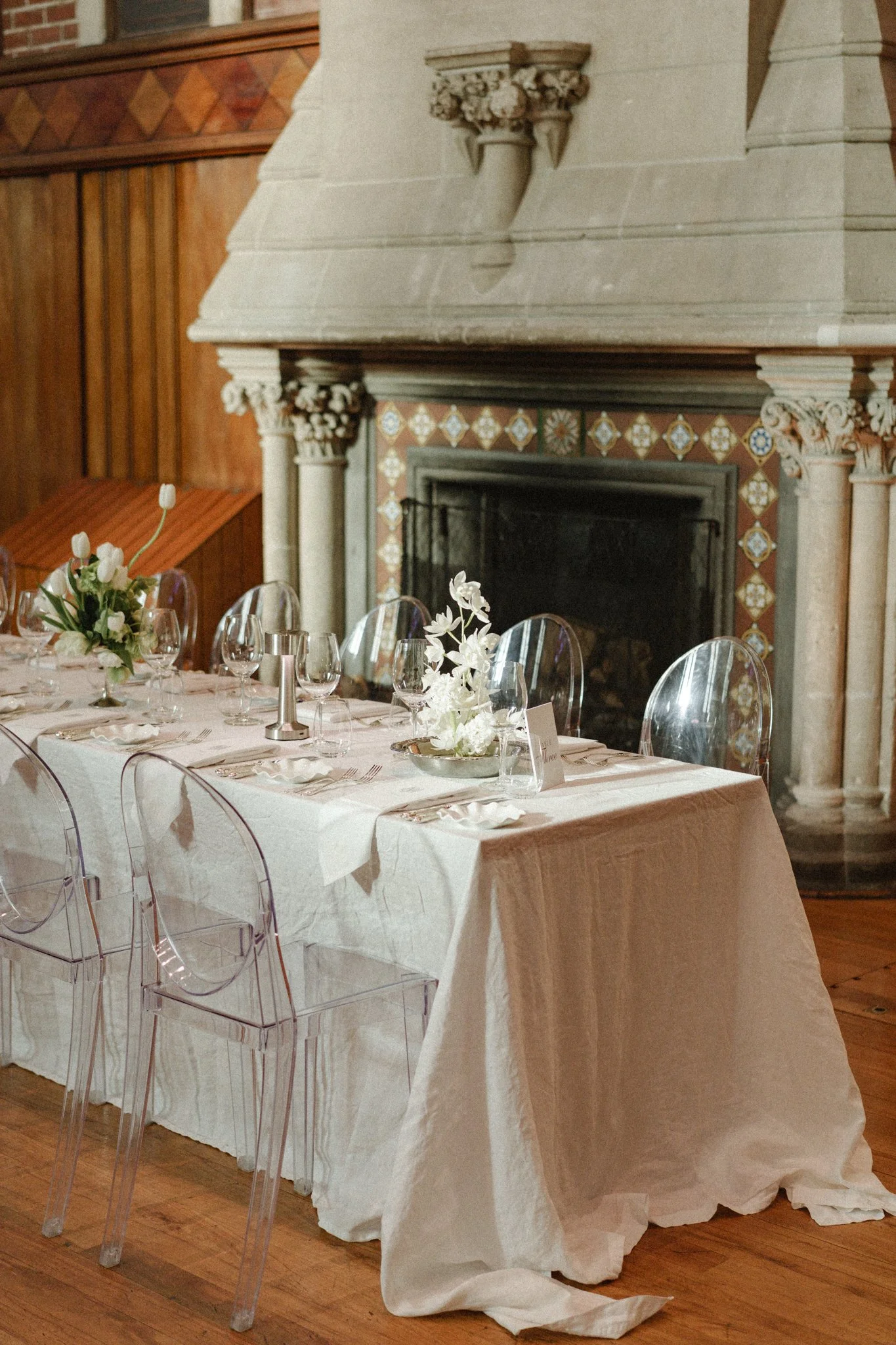 Wedding reception table with white linen and flowers beside an ornate fireplace.