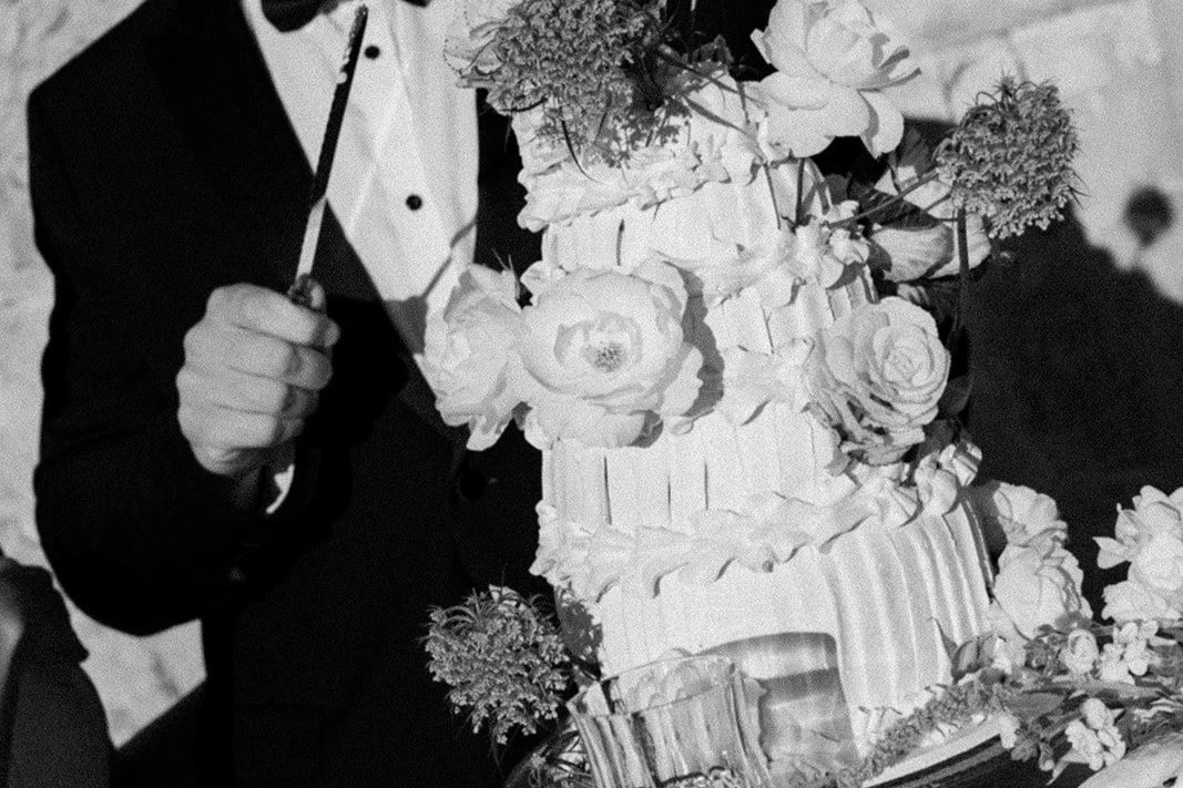 A person holding a cake knife near a multi-tiered wedding or celebration cake decorated with flowers.