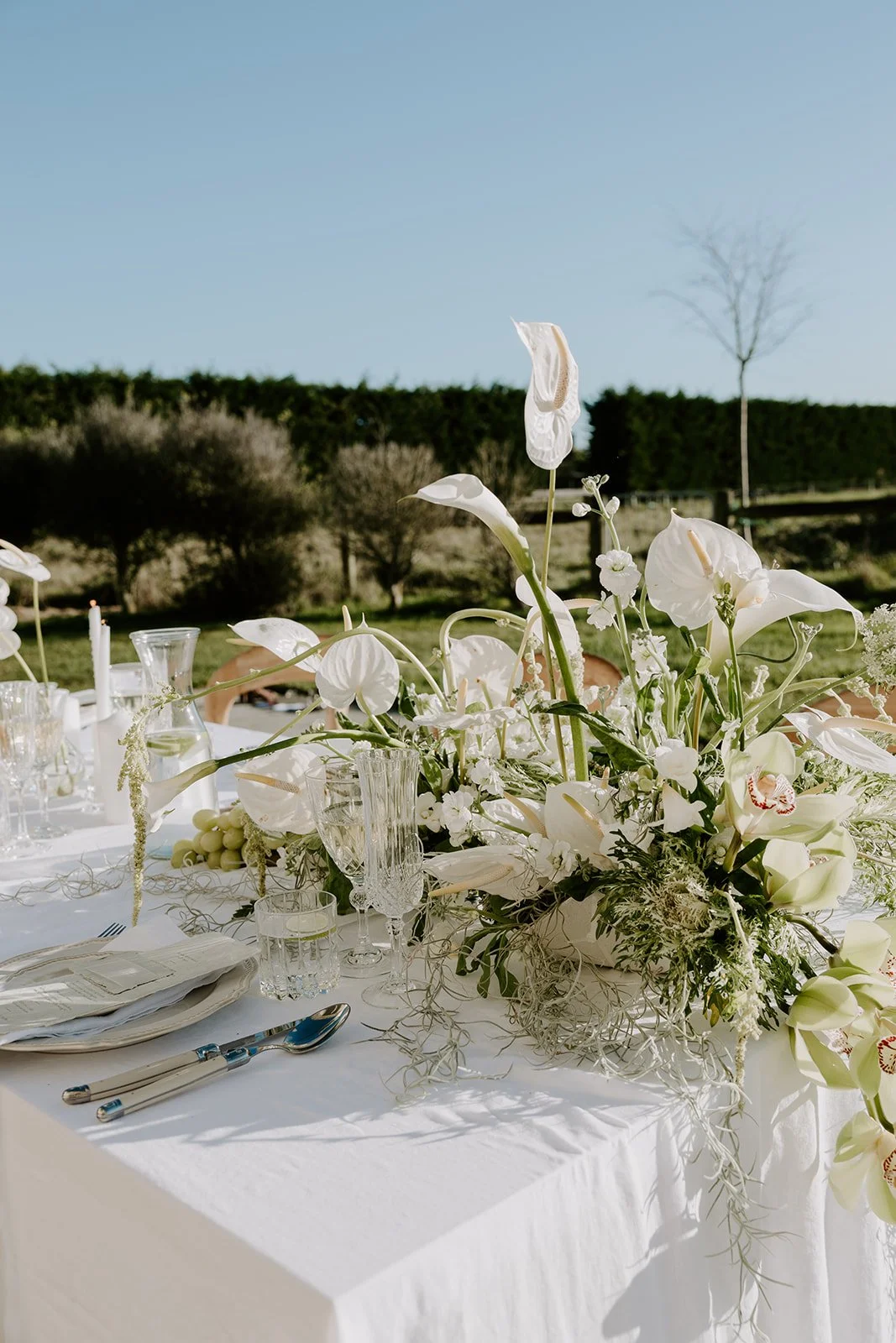 A white floral centerpiece on a table set for an outdoor event, with glassware, plates, and silverware.
