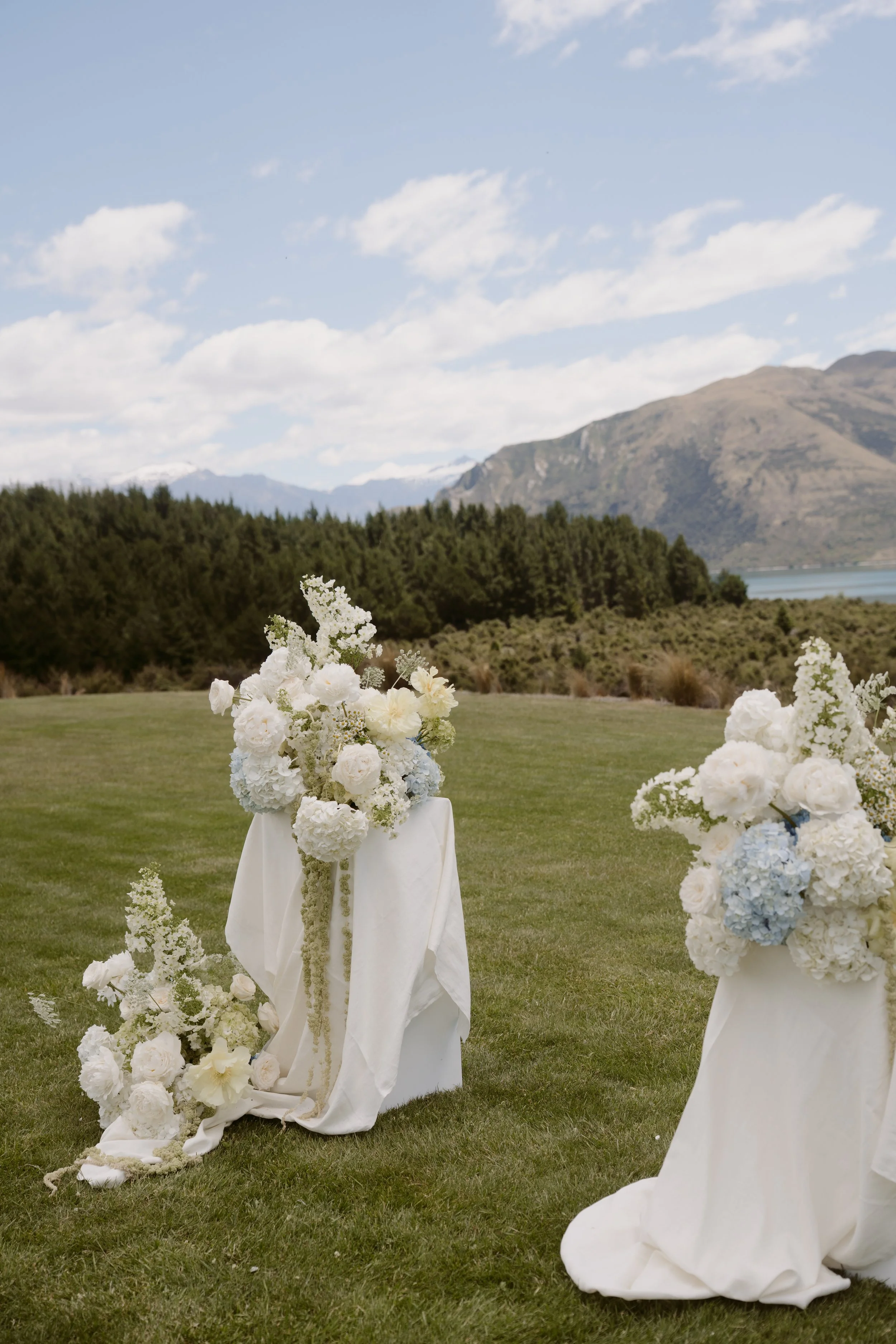 Outdoor wedding setup with floral arrangements on white stands, set against a scenic backdrop of mountains, a lake, and a partly cloudy sky.