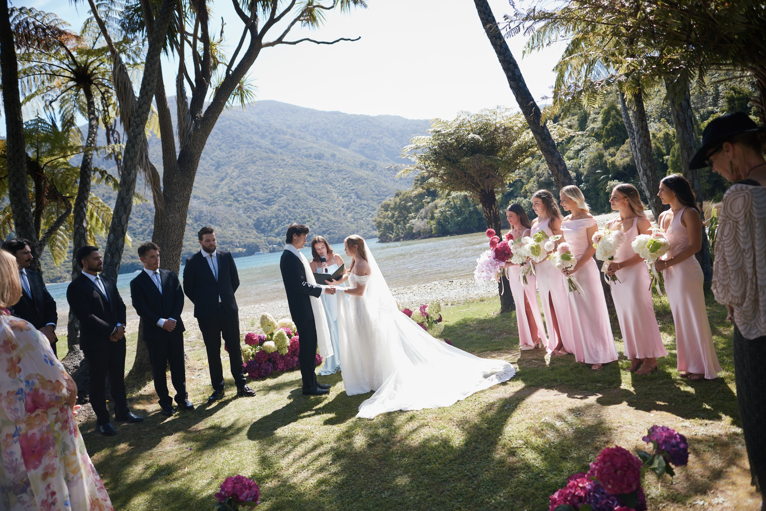 A wedding ceremony outdoors by a body of water with mountains in the background, featuring a bride and groom exchanging vows, surrounded by bridesmaids in pink dresses holding bouquets, and groomsmen in dark suits.