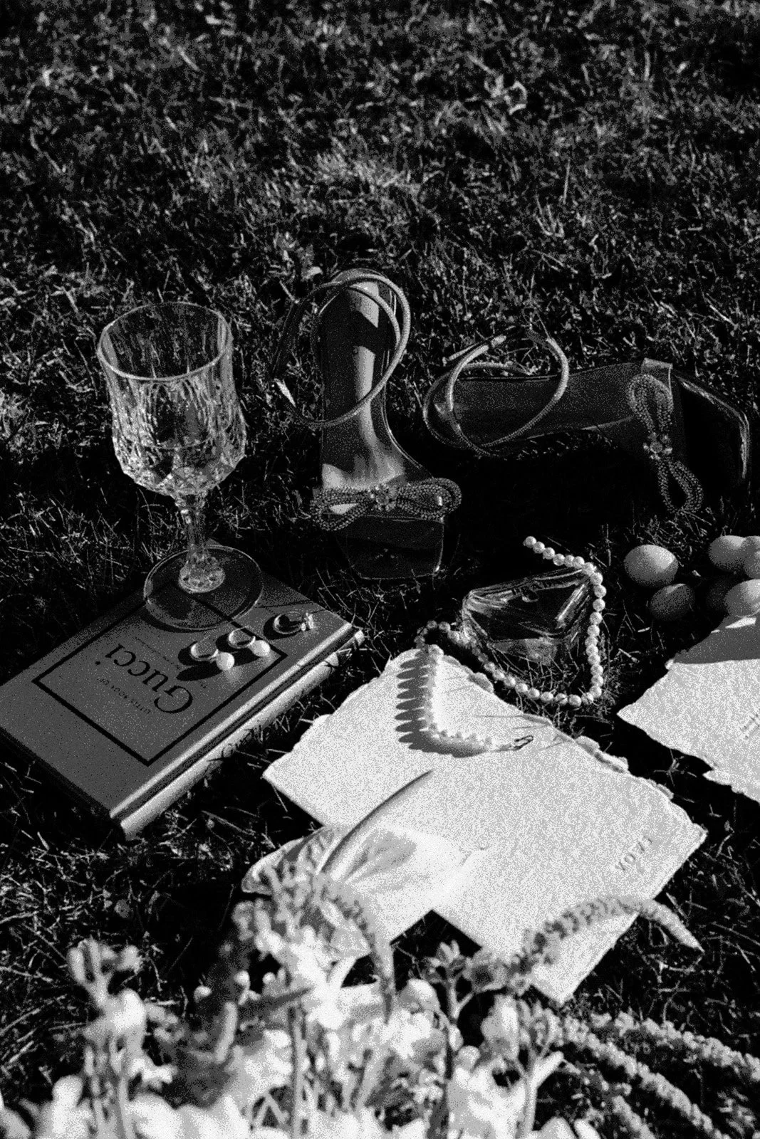 Wedding accessories, embroidered fabric, silverware and glassware arranged on grass, in black and white.