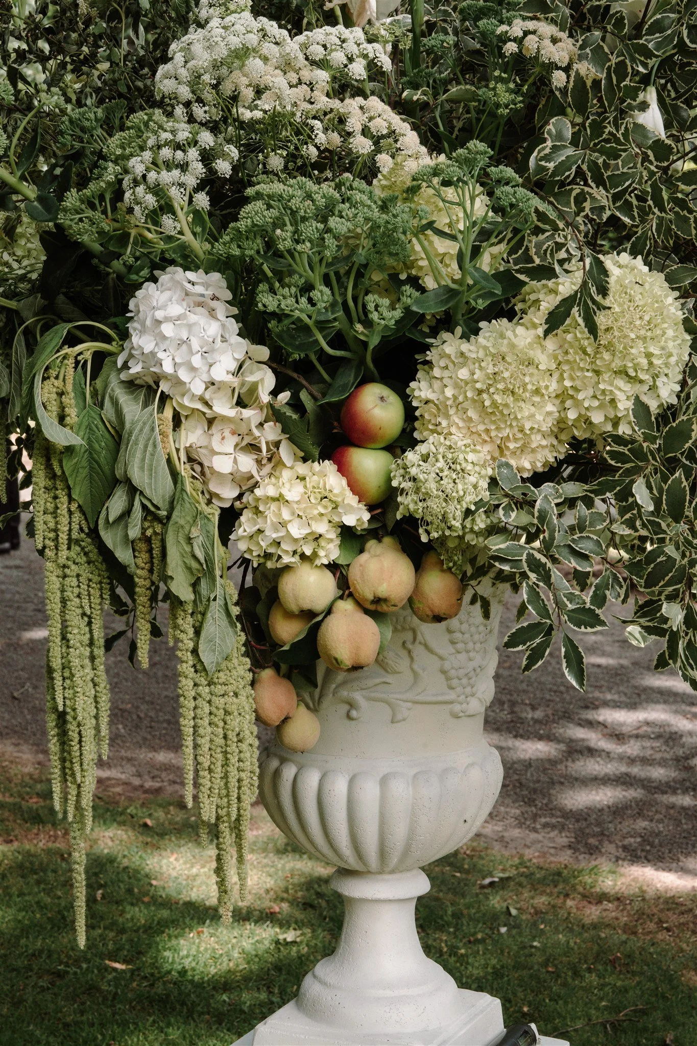 Wedding floral arrangement with green hydrangeas, trailing foliage and fruit in a stone urn.