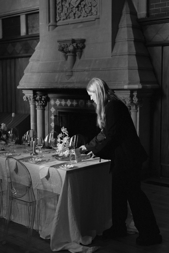 Woman in dark clothing decorating a dining table in a room with a large stone fireplace and ornate woodwork, in black and white.