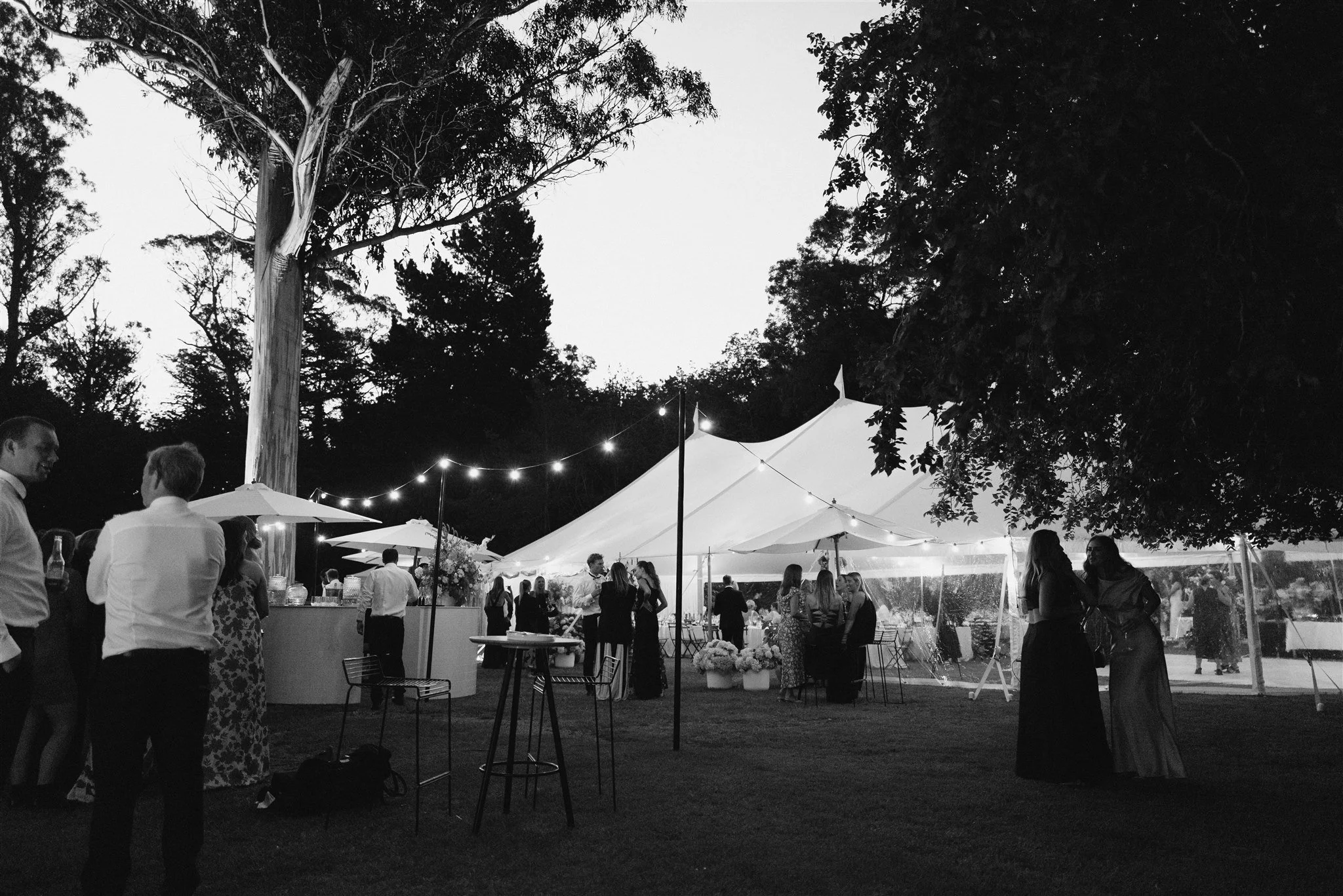 Guests gathering outside a wedding marquee beneath festoon lights, in black and white.