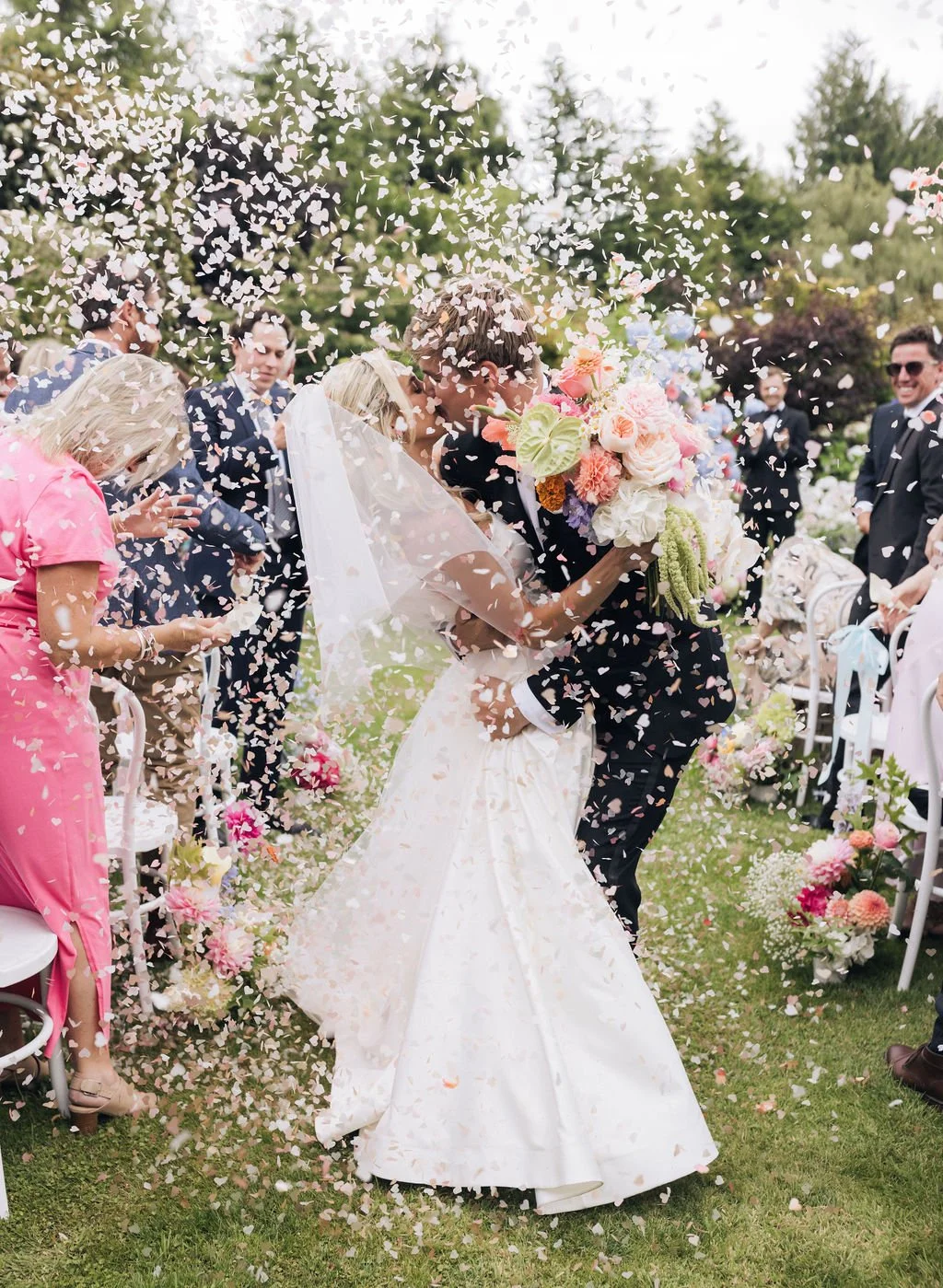 Bride and groom sharing a kiss during their wedding reception, surrounded by guests and showered with flower petals.