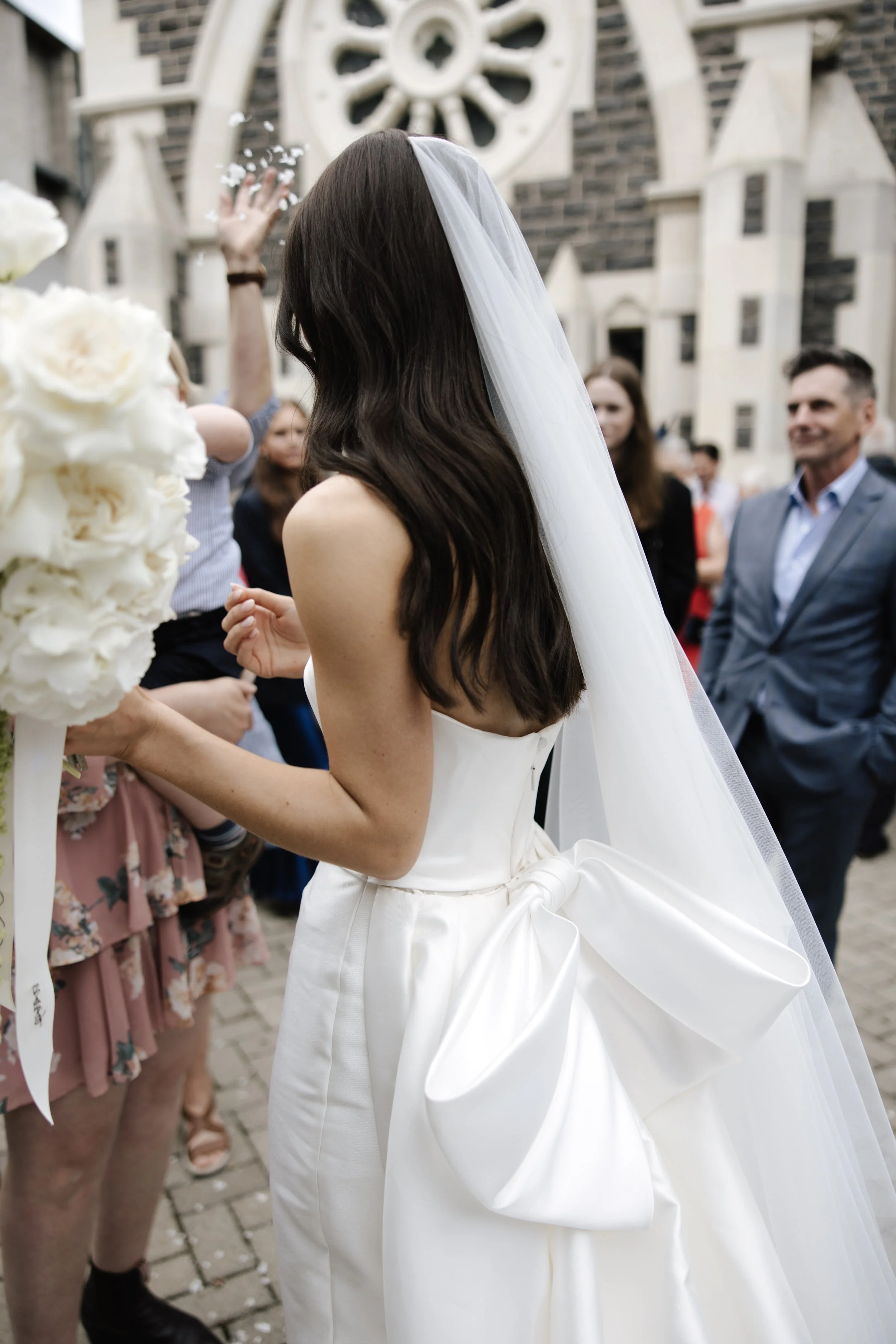 Bride with a large satin bow and veil greeting guests outside a stone church.