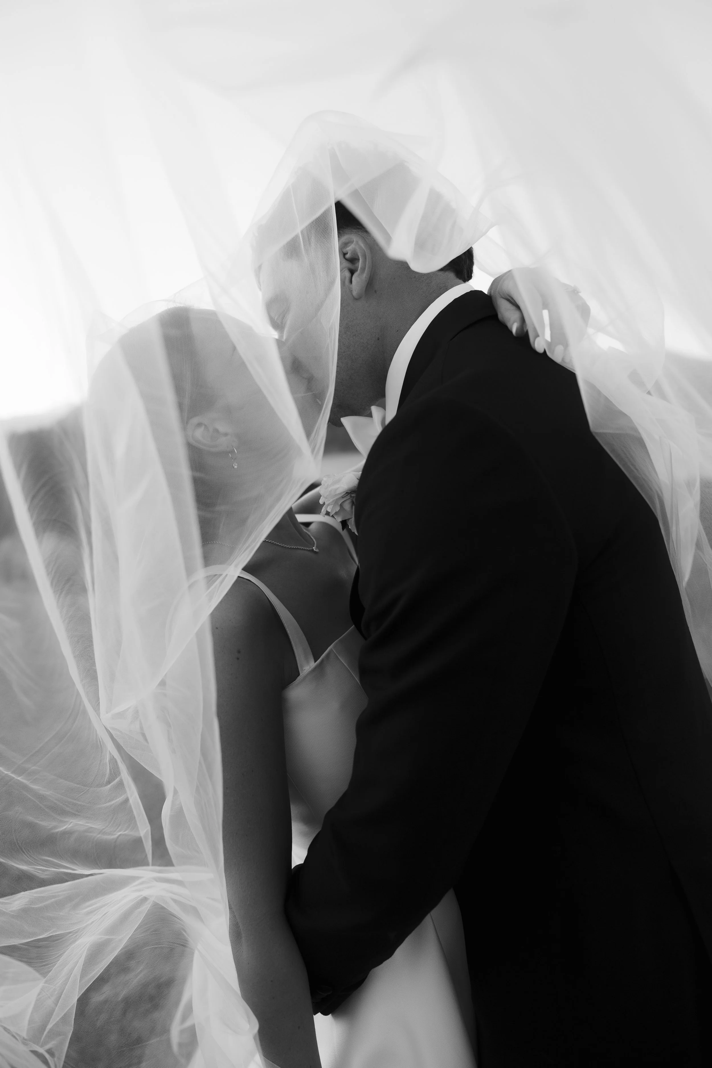 A groom and bride share a kiss under a veil at their wedding, with formal attire and delicate tulle fabric surrounding them.
