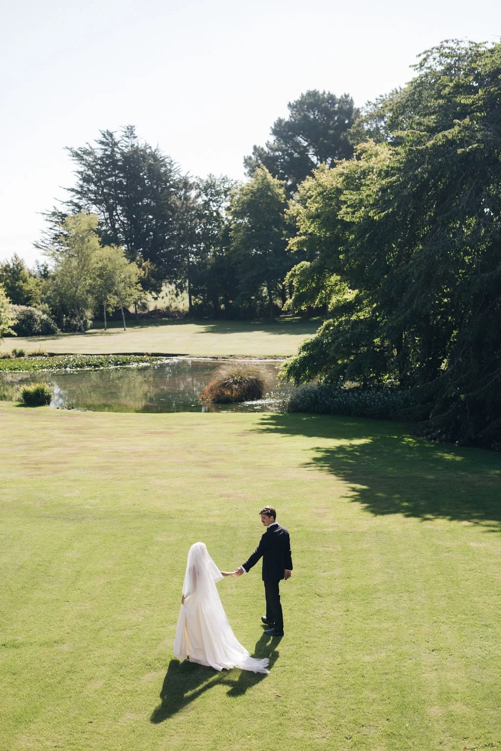 Newlyweds walking hand in hand across a garden lawn beside a pond.