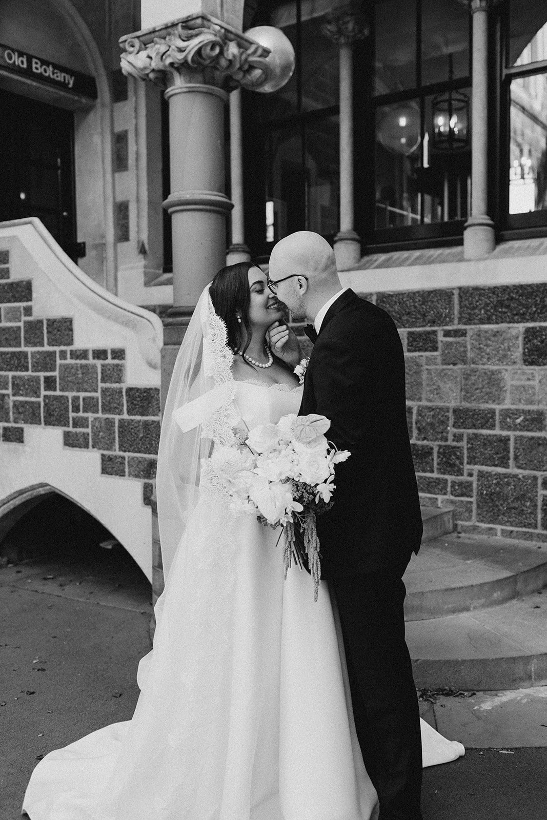 A black and white photo of a bride and groom standing outside in front of a building with stone and brick details, sharing a close moment with their foreheads touching.