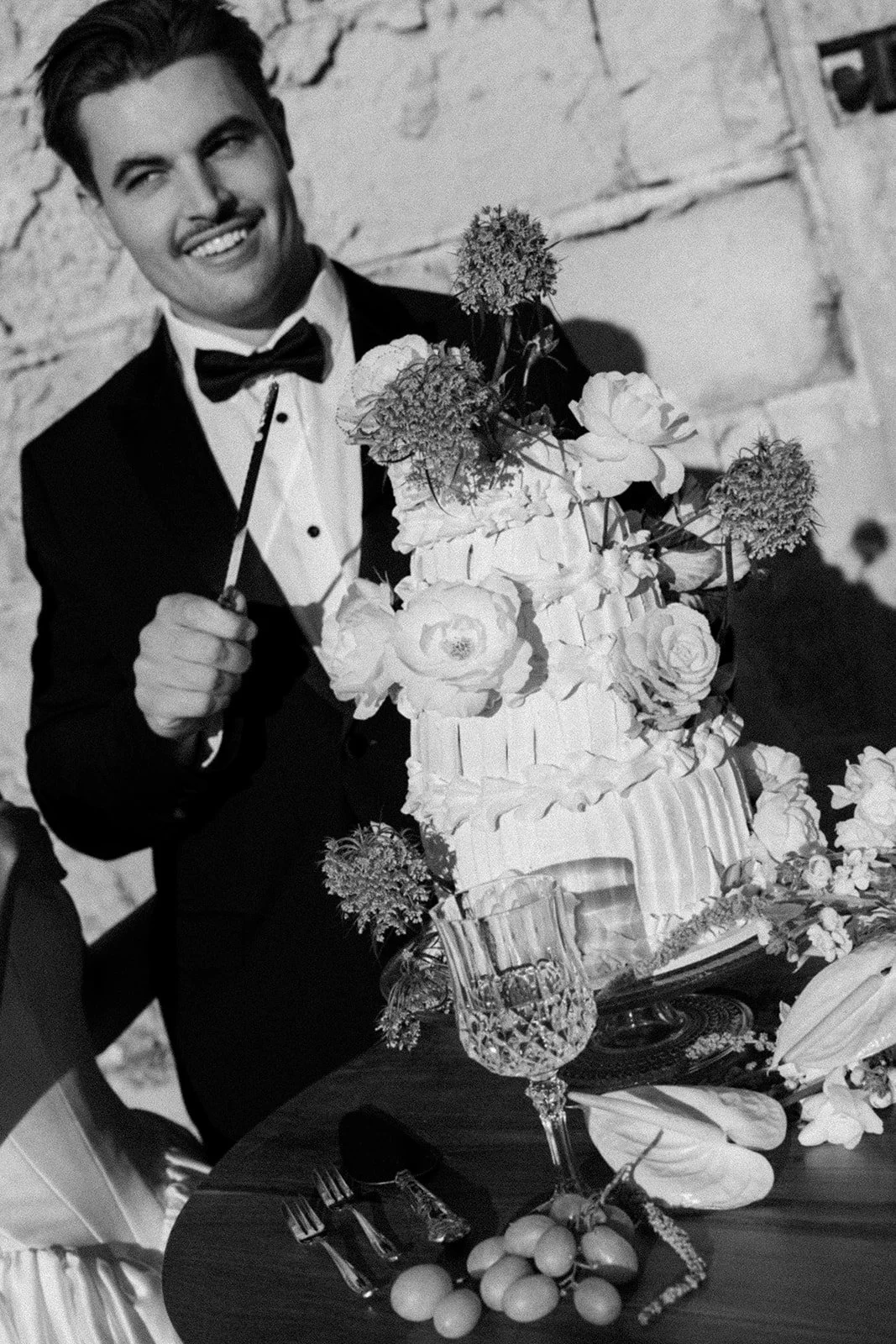 A man in a tuxedo holding a knife, standing next to a large decorated cake with flowers, on a table with wine glasses, cutlery, and fruit, at a celebration or wedding reception.