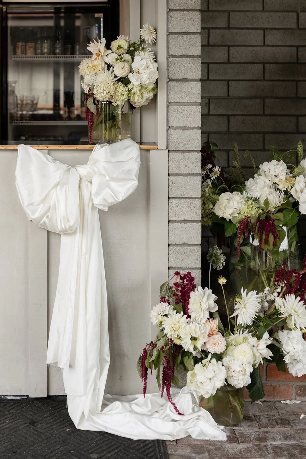 White wedding floral installation with draped fabric and a large bow beside a brick wall.