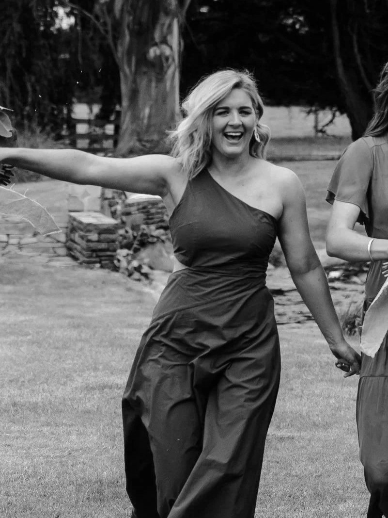 A woman in a one-shoulder dress smiling and holding hands with a person outside, with trees and outdoor stones in the background.