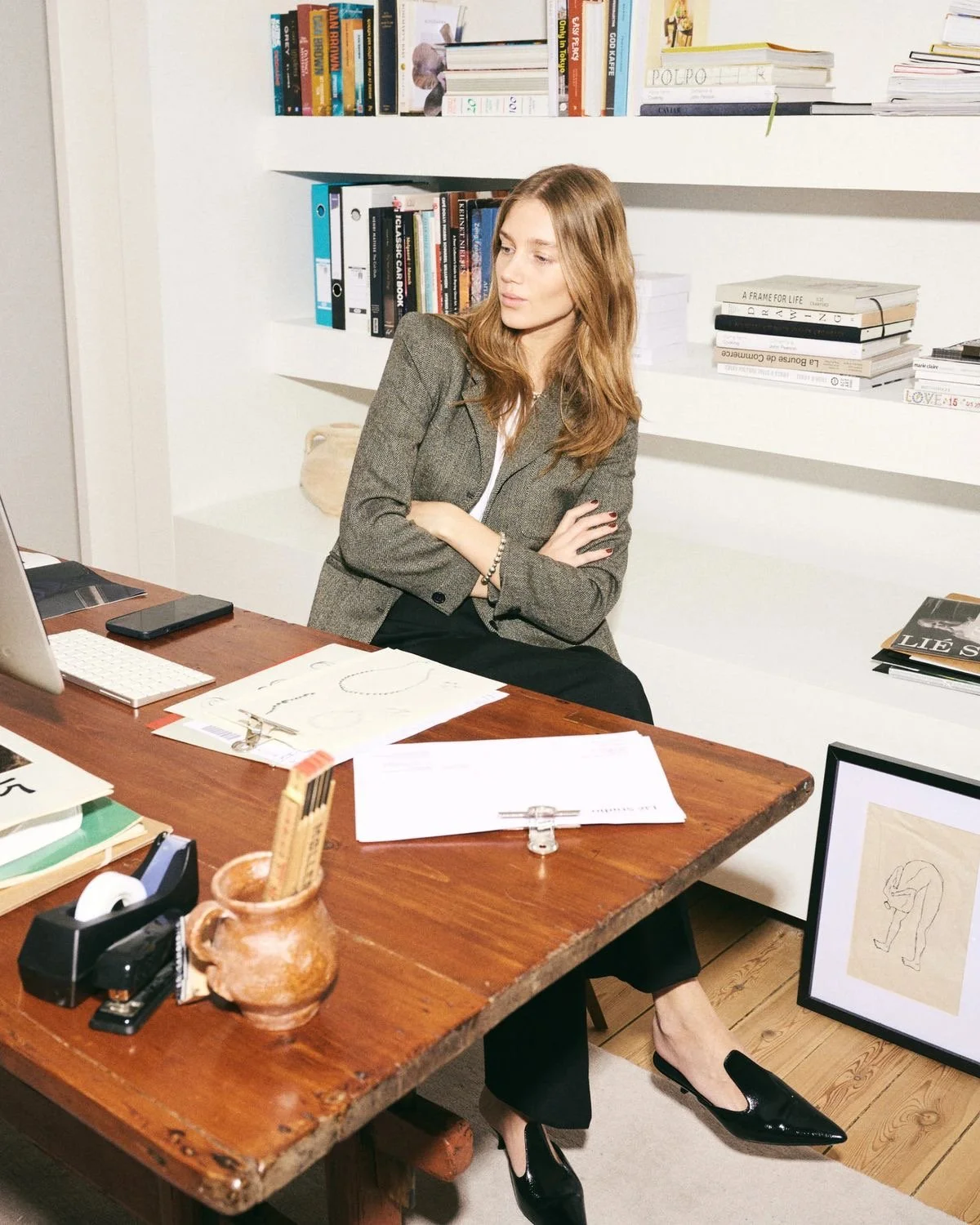 A woman with reddish hair sitting at a cluttered wooden desk in an office or study, with a bookshelf behind her filled with books and stacks of paper, and a framed drawing of a person bending over on the floor near her feet.