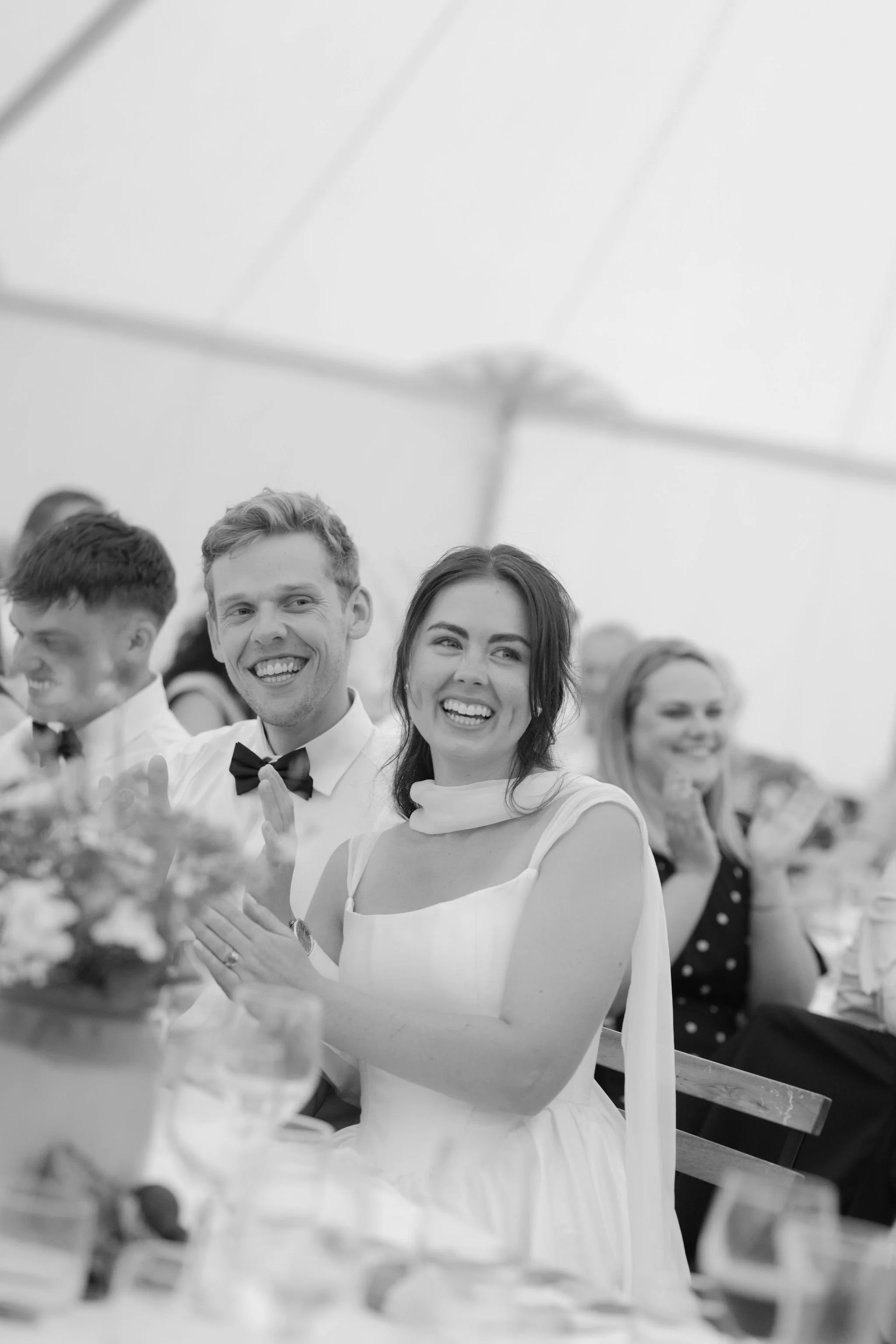 People attending a formal event, smiling and clapping, seated with tables and plates, in a large tent or similar structure.