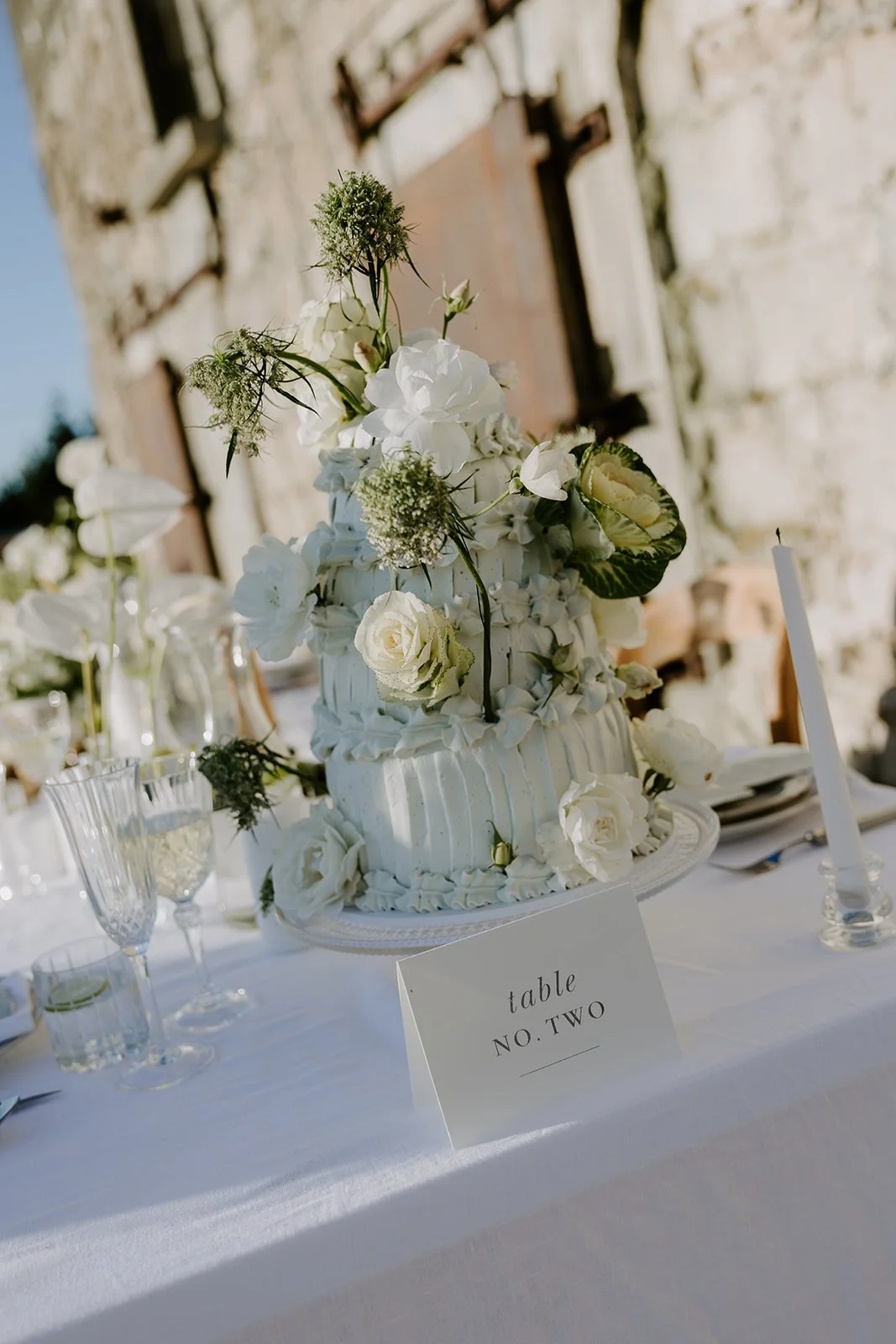 A tall wedding cake decorated with white flowers and greenery, placed on a table with glassware and a table card labeled "Table No. Two." In the background, a stone wall and outdoor setting.