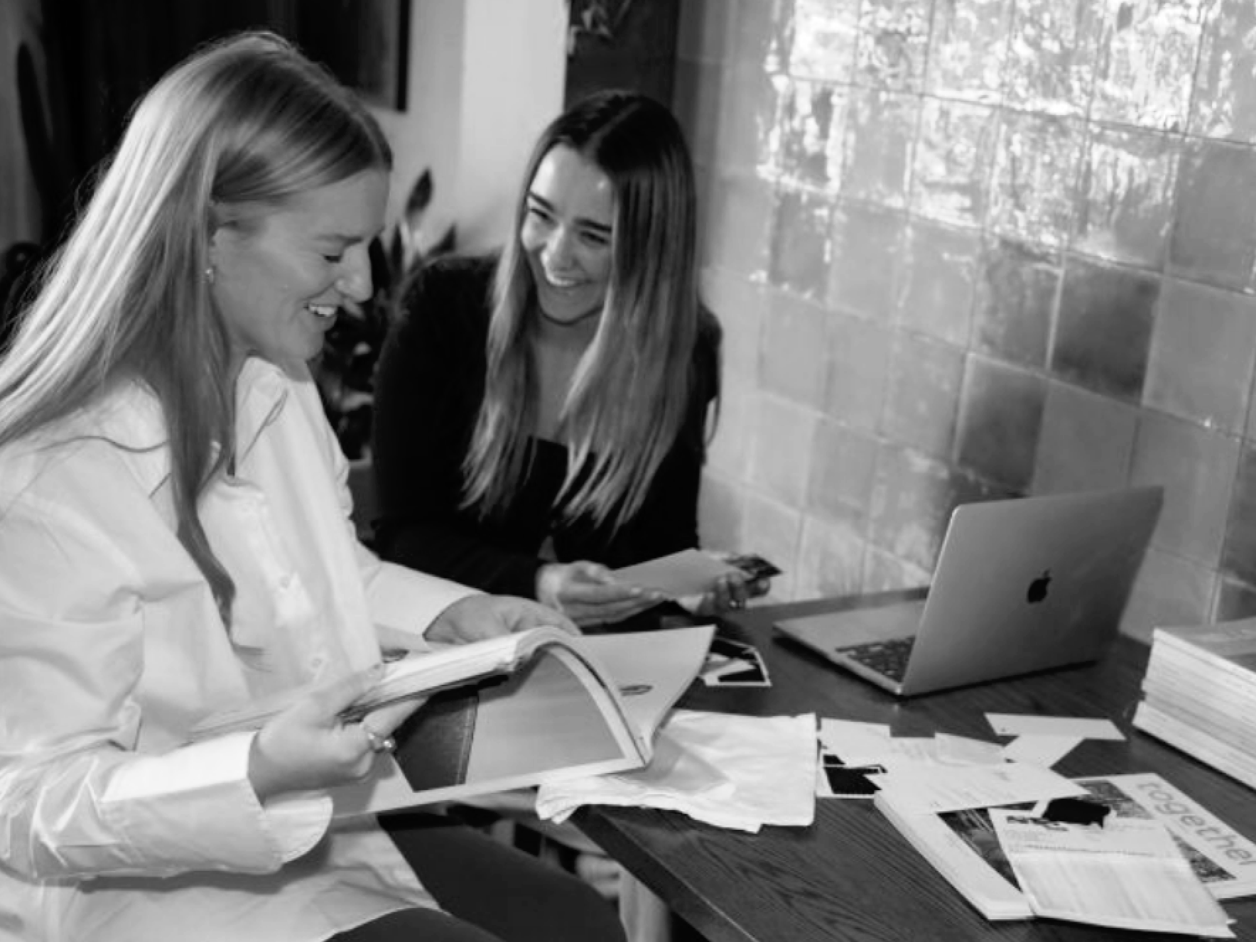 Two women sitting at a table with papers, notebooks, and a laptop, engaged in work and smiling.