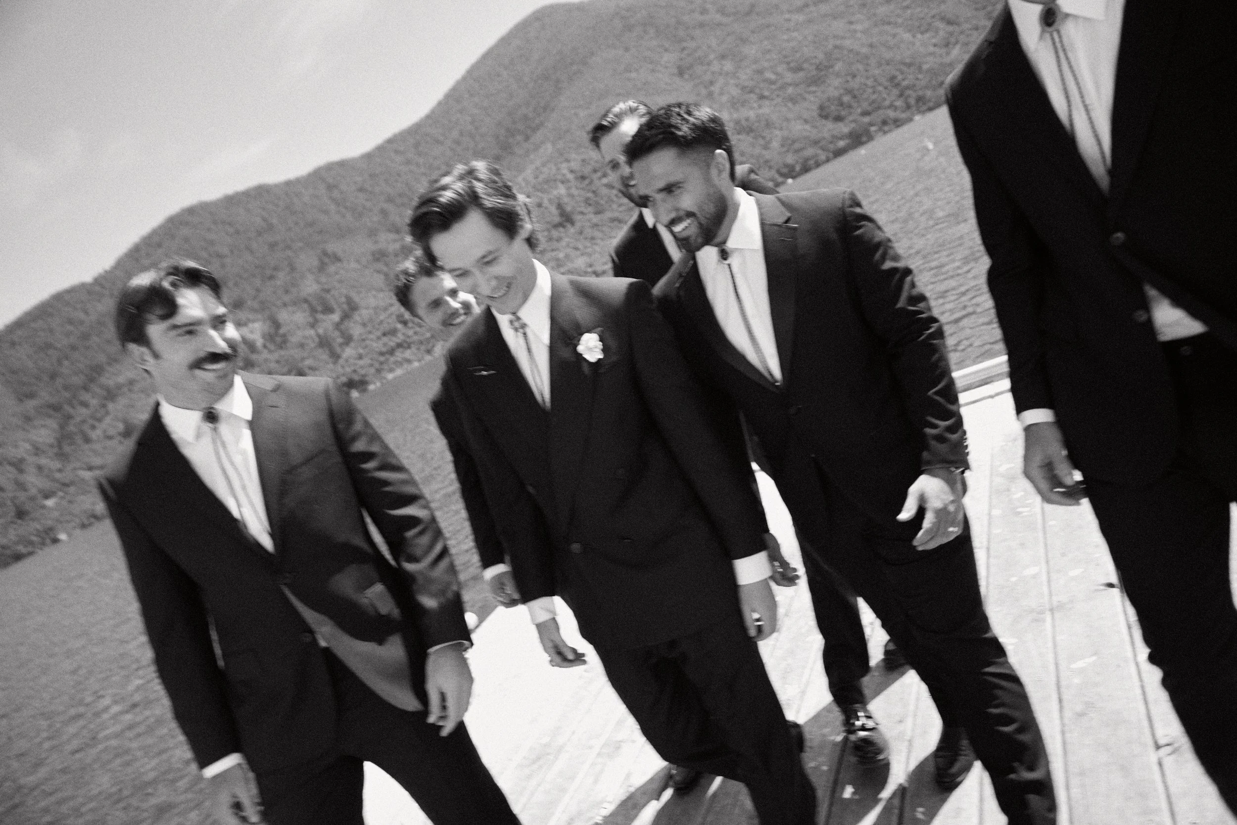 Group of five men dressed in formal suits holding hands and walking outdoors near a body of water with mountains in the background, smiling and laughing together.