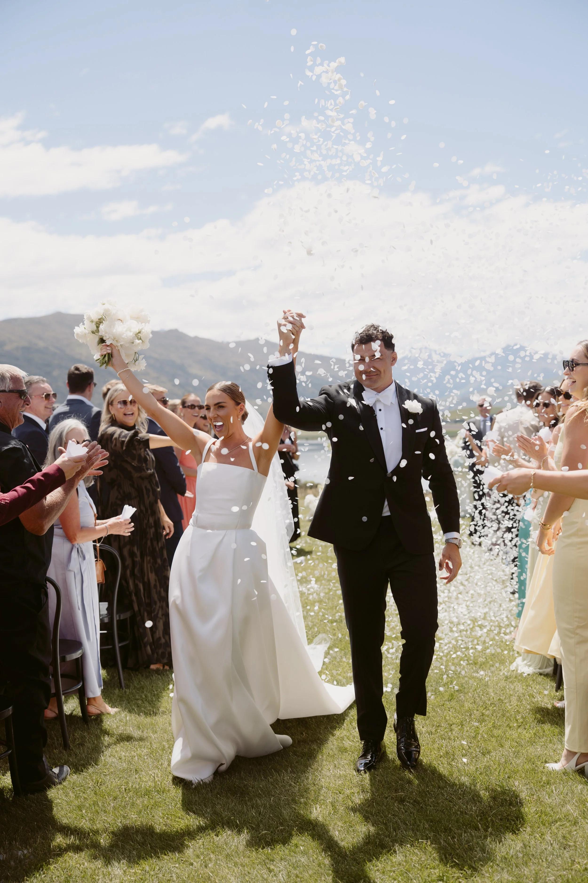 A bride and groom celebrate with confetti during their outdoor wedding ceremony, surrounded by seated guests and scenic mountains in the background.