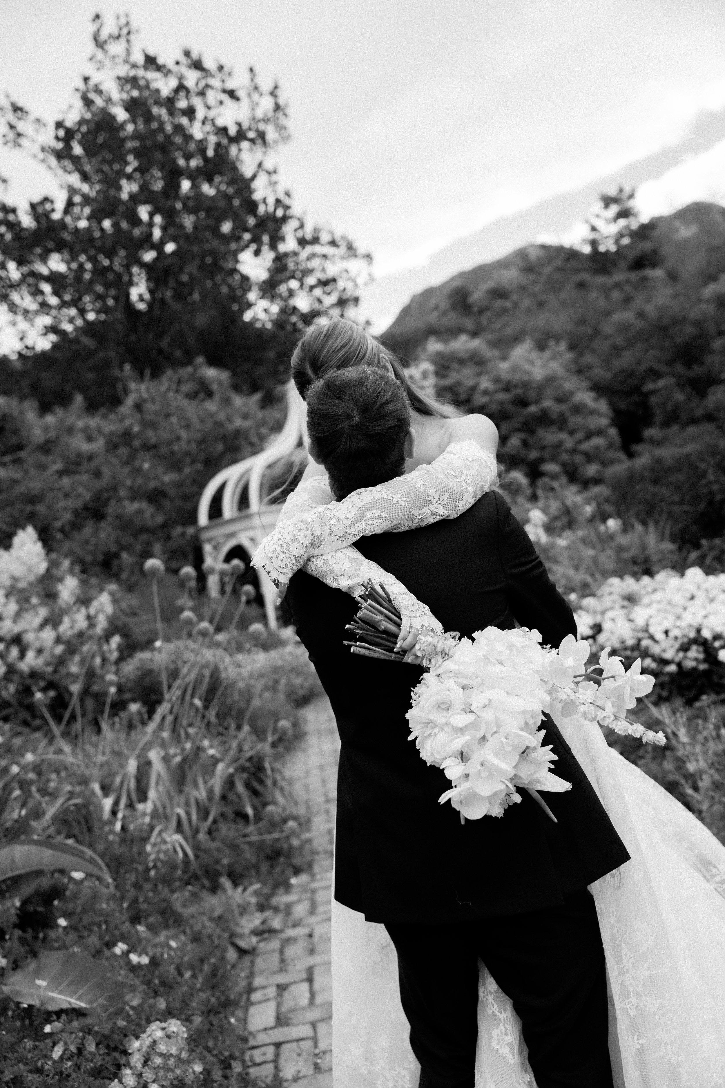 A person in formal attire holding a woman in a lace dress in an outdoor garden setting, with trees and hills in the background.