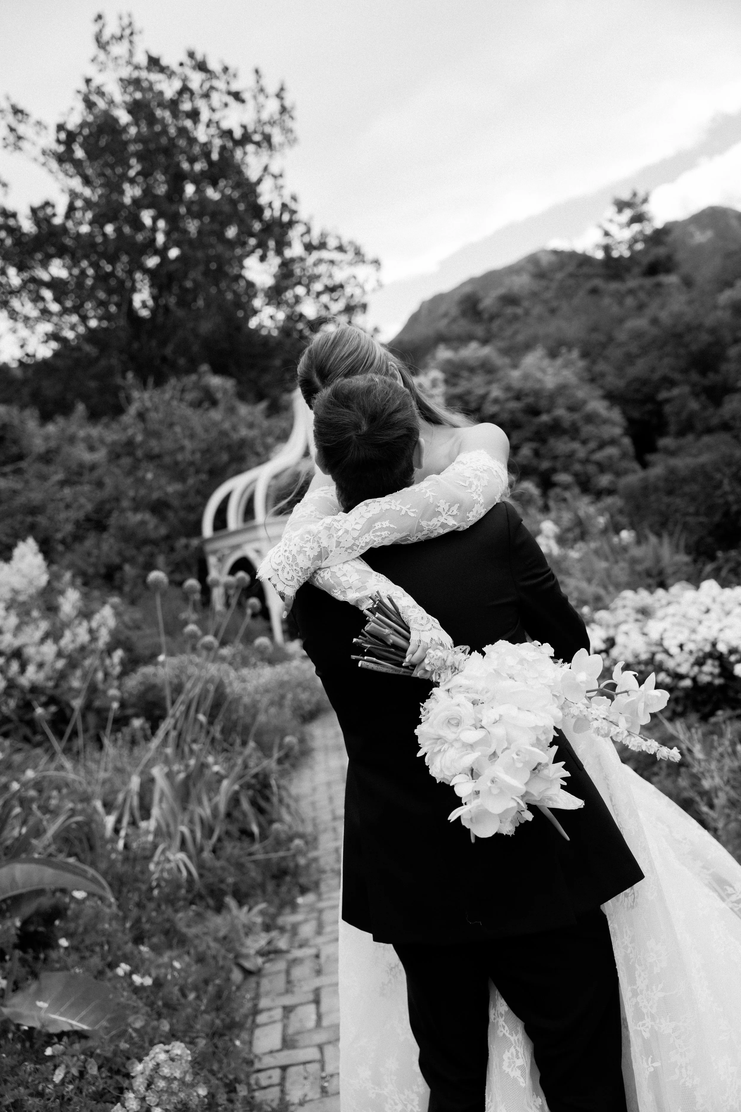 Newlyweds embracing on a garden path, in black and white.