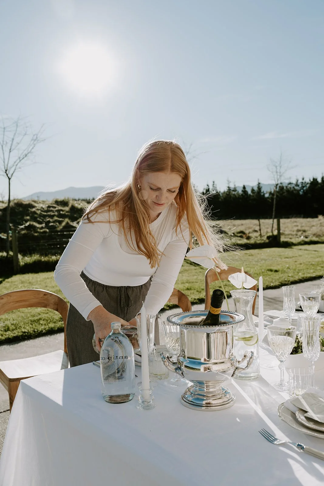 Wedding stylist arranging glassware on a sunlit outdoor reception table.