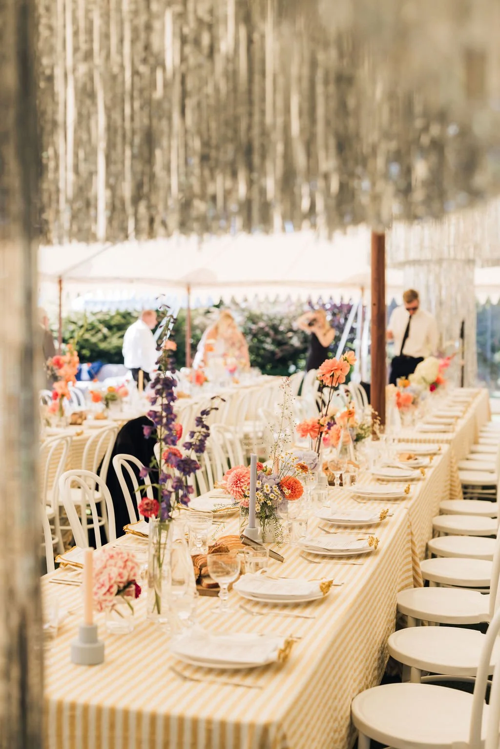 A long dining table set for a celebration outdoors, decorated with colorful floral arrangements, candles, and tableware, under a covered canopy with people in the background.