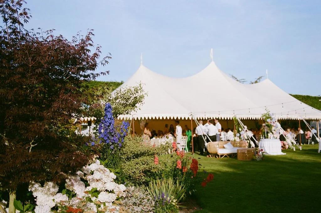 A large white tent set up outdoors amid a garden of colorful flowers, with people gathered underneath for an event.