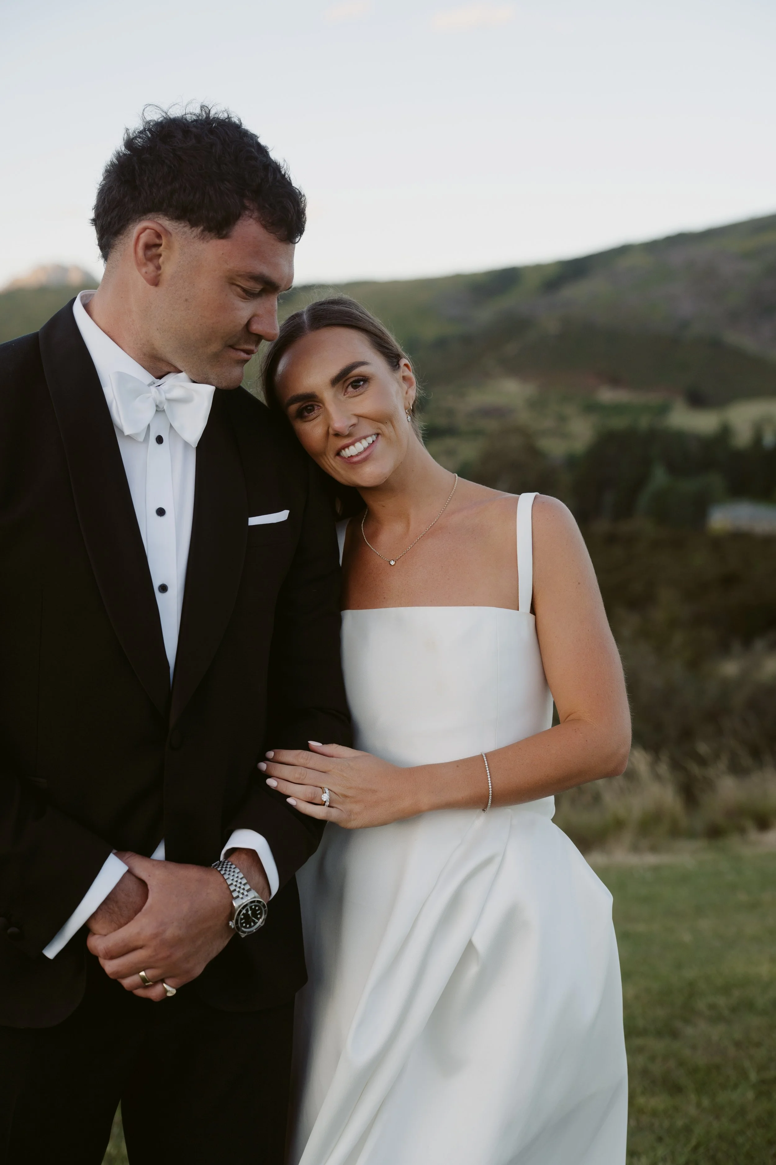 A newlywed couple dressed in formal wedding attire outdoors with a backdrop of green hills and sky. The man is in a black tuxedo with a white bow tie, and the woman is in a white strapless wedding gown, smiling and leaning her head on his shoulder.