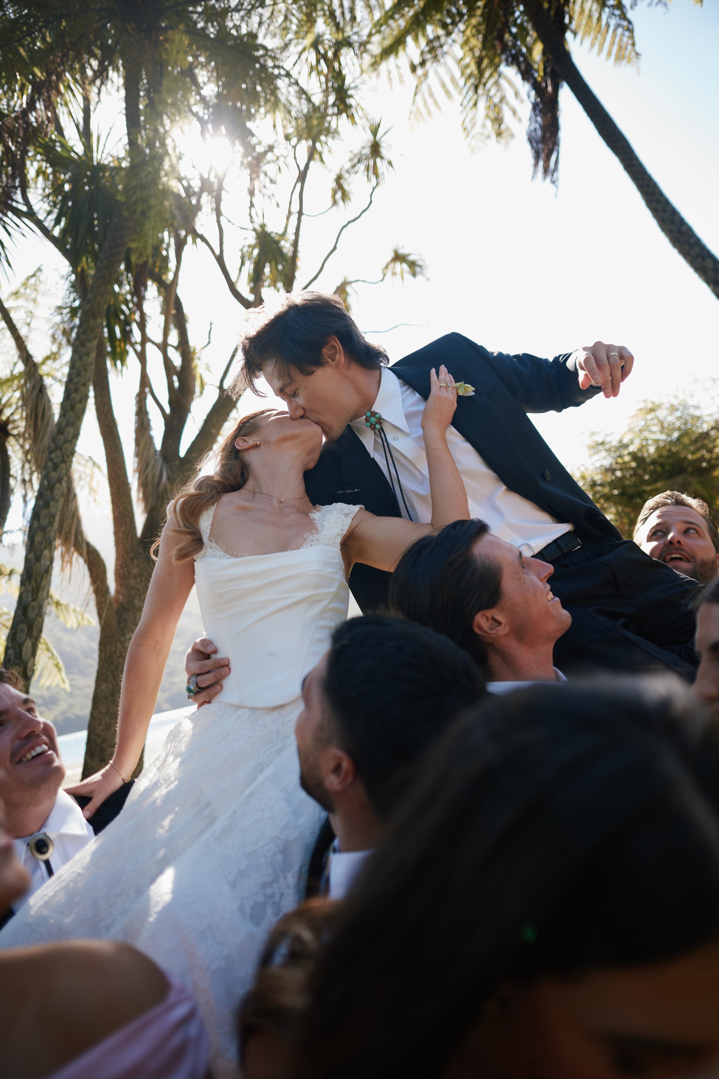 A couple kissing during a wedding celebration outdoors, surrounded by friends, with palm trees and sunlight in the background.